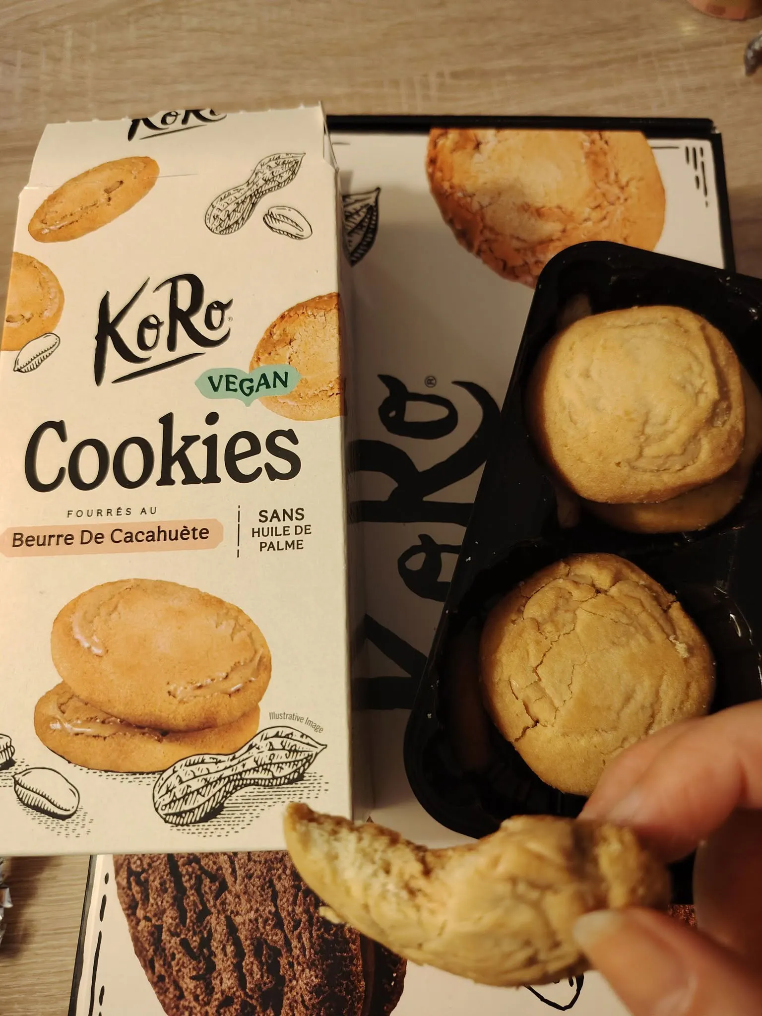 eine geöffnete packung koro vegane erdnussbutter-kekse neben einer schwarzen kunststoffschale mit weiteren keksen, wobei eine hand einen angebrochenen keks hält.