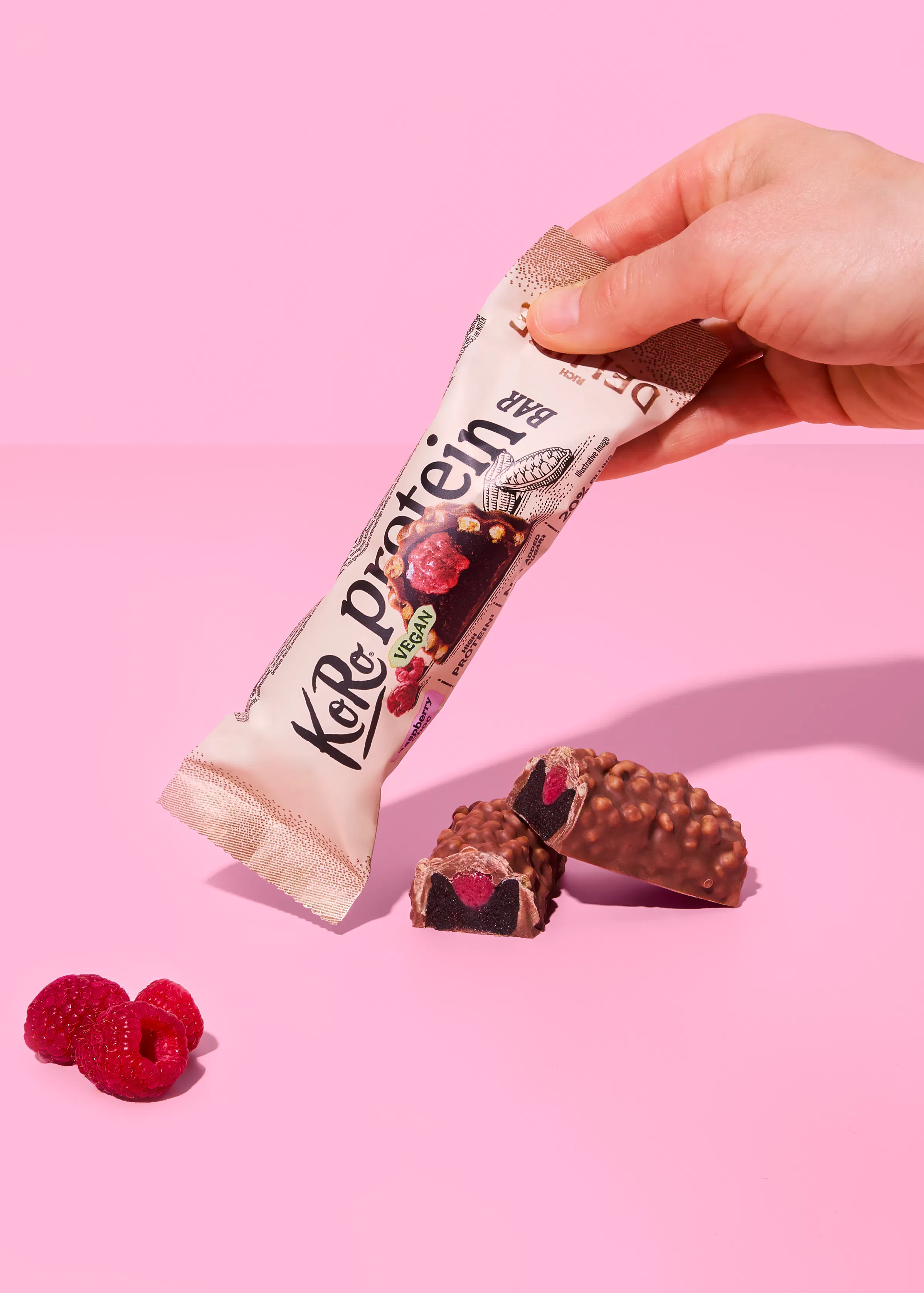 a hand holds a koko protein bar with raspberry filling, next to two broken bar pieces and fresh raspberries on a pink background.