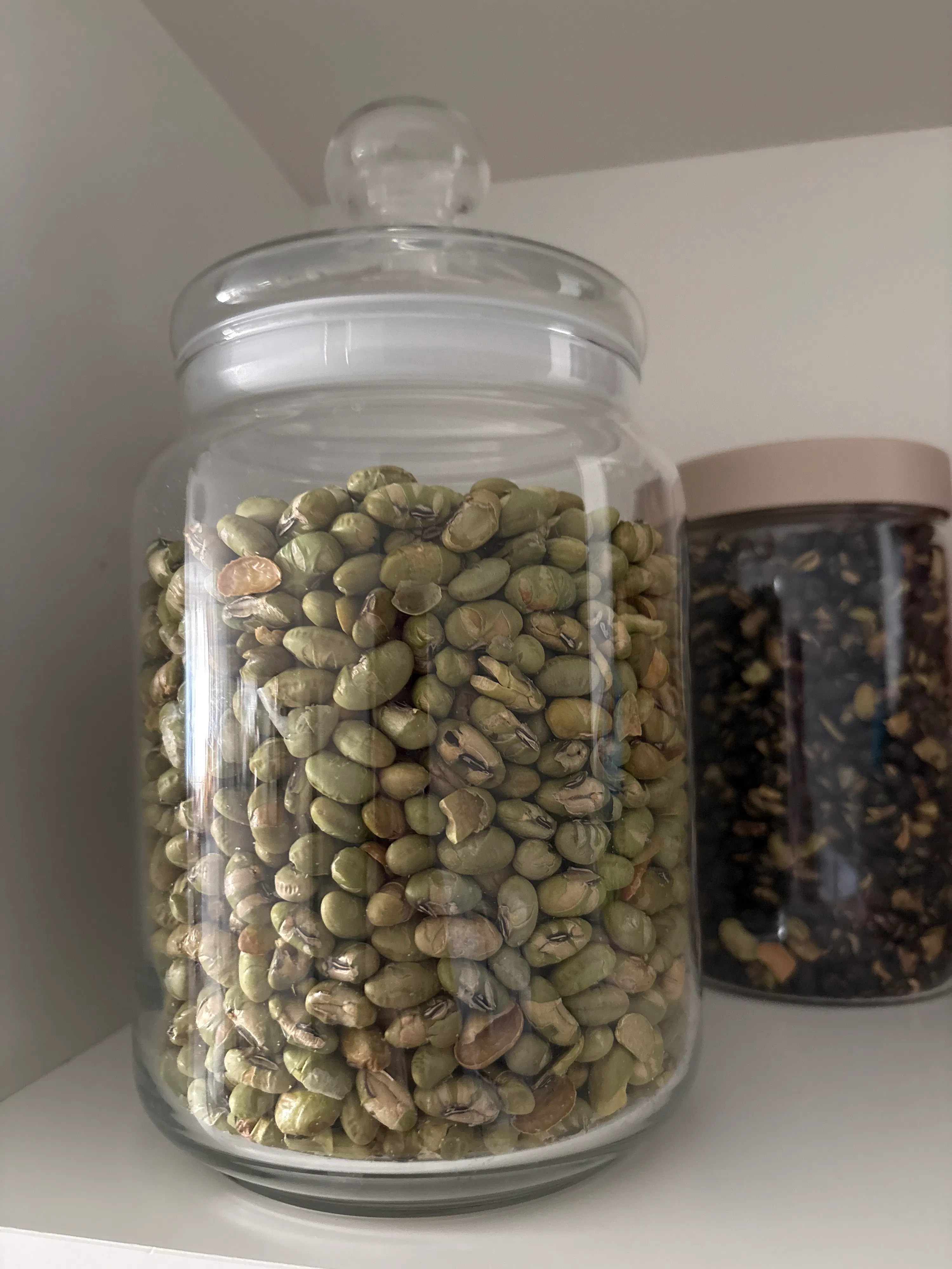un barattolo di vetro trasparente pieno di fagioli edamame verdi su uno scaffale bianco.