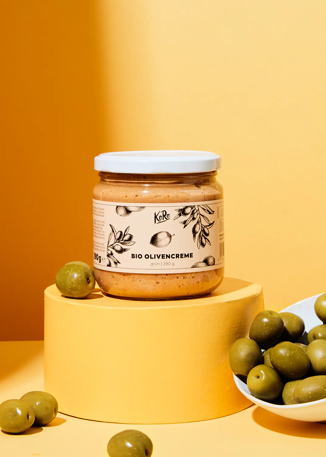 un bocal de creme d' olives bio est assis sur un support jaune entouré d' olives