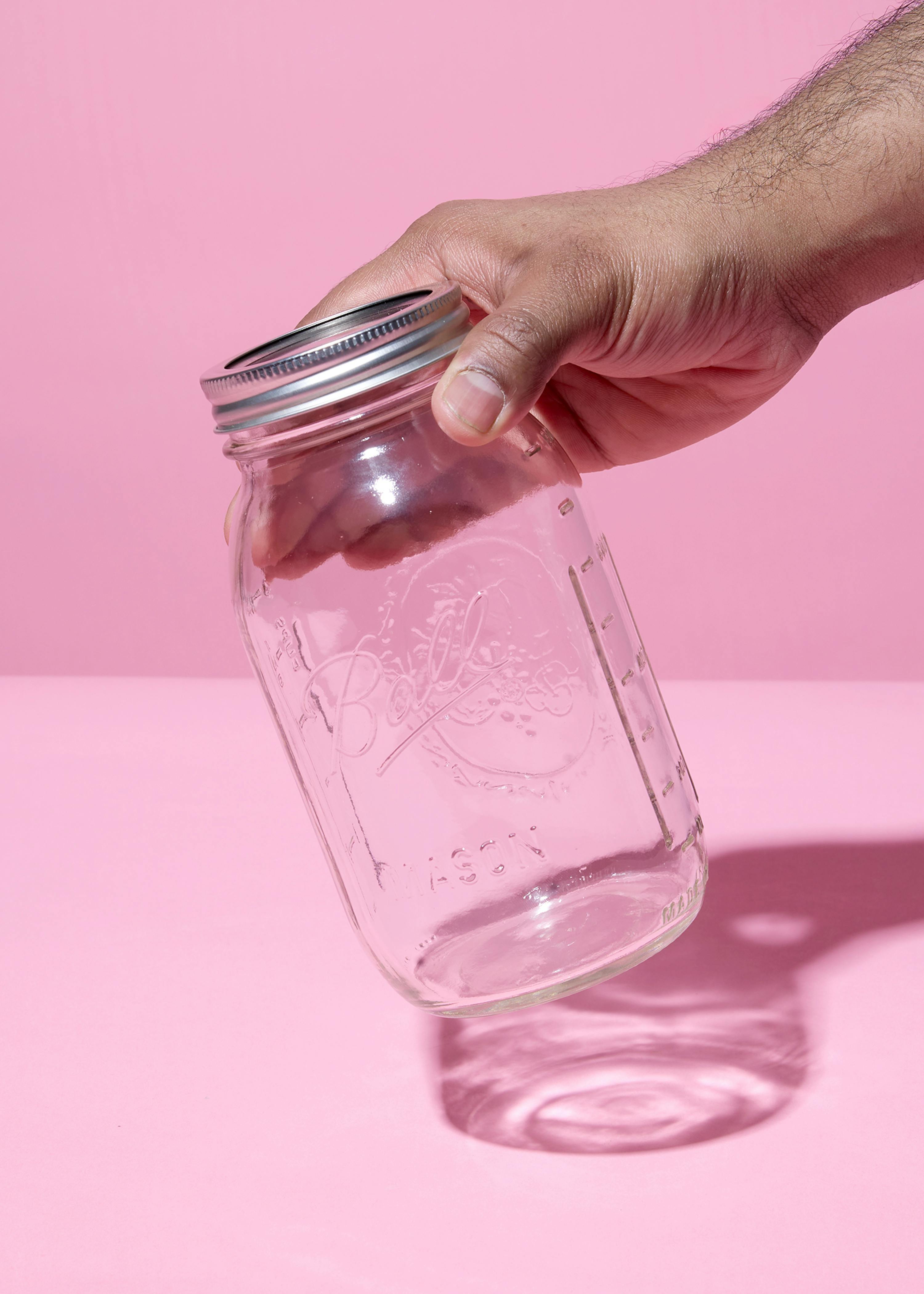a dark-skinned hand holds a clear glass mason jar with a silver lid against a pink background.