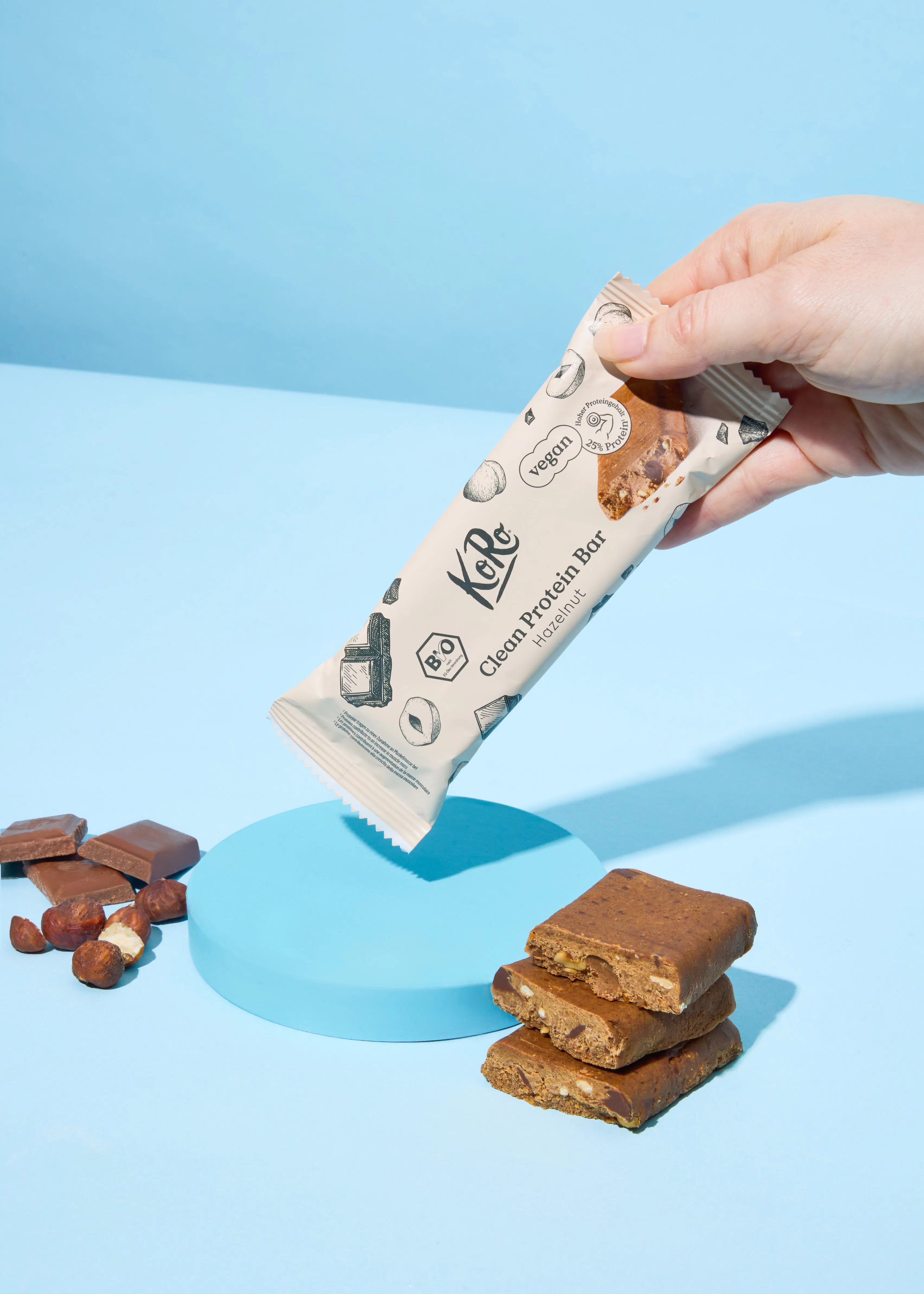 une main tient une barre protéinée koro saveur noisette, avec des morceaux de barre coupés, des noisettes et des carrés de chocolat sur un fond bleu.
