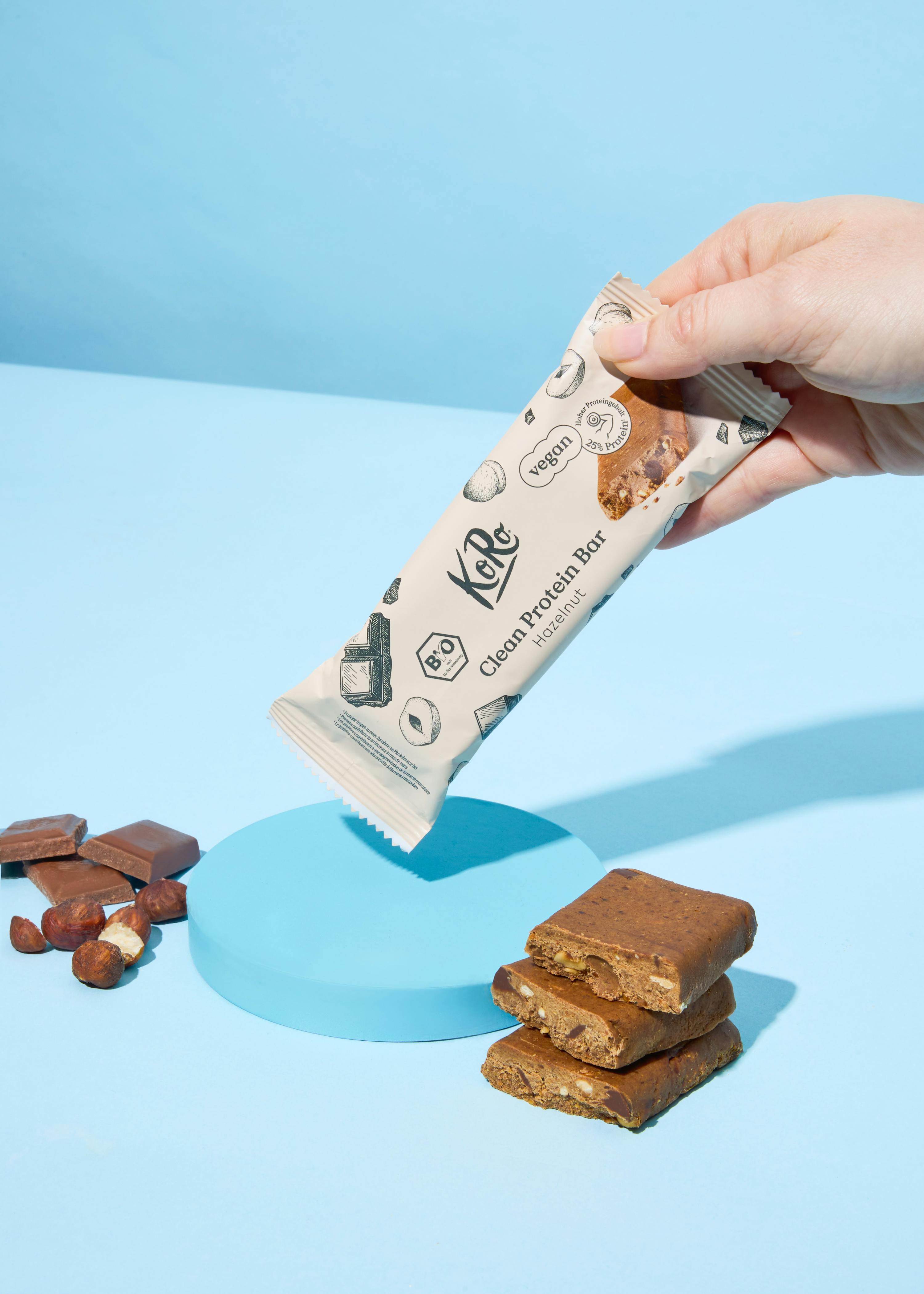 eine hand hält eine verpackung des koro clean protein riegels haselnuss, während daneben auf einem hellblauen hintergrund geschnittene riegel, haselnüsse und schokoladenstücke liegen.