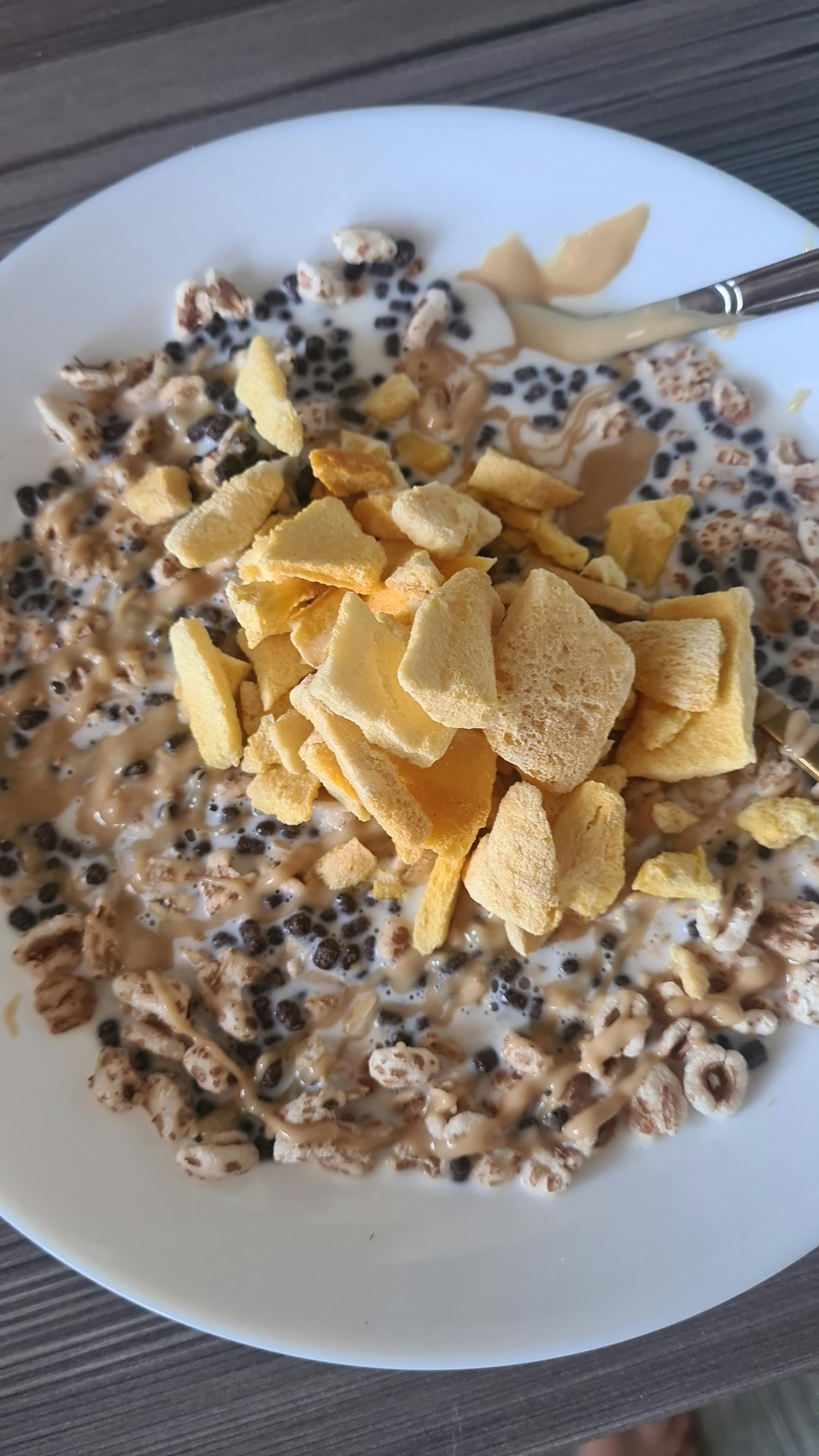 una ciotola di cereali con latte, palline di cioccolato, cereali soffiati, burro di arachidi e pezzi gialli di frutta essiccata.