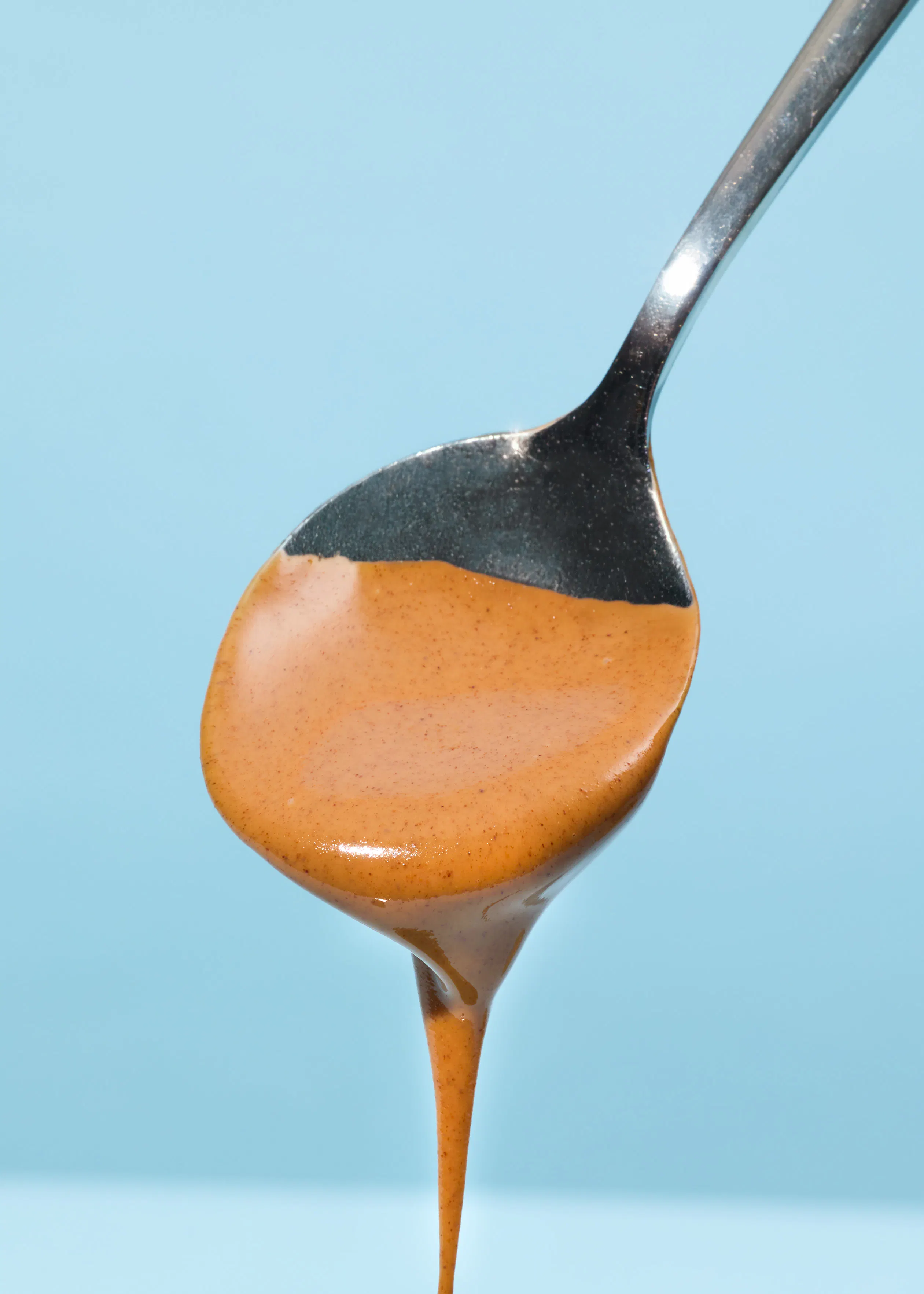 un cucchiaio d'argento pieno di crema spalmabile marrone chiaro che cola, su uno sfondo blu.