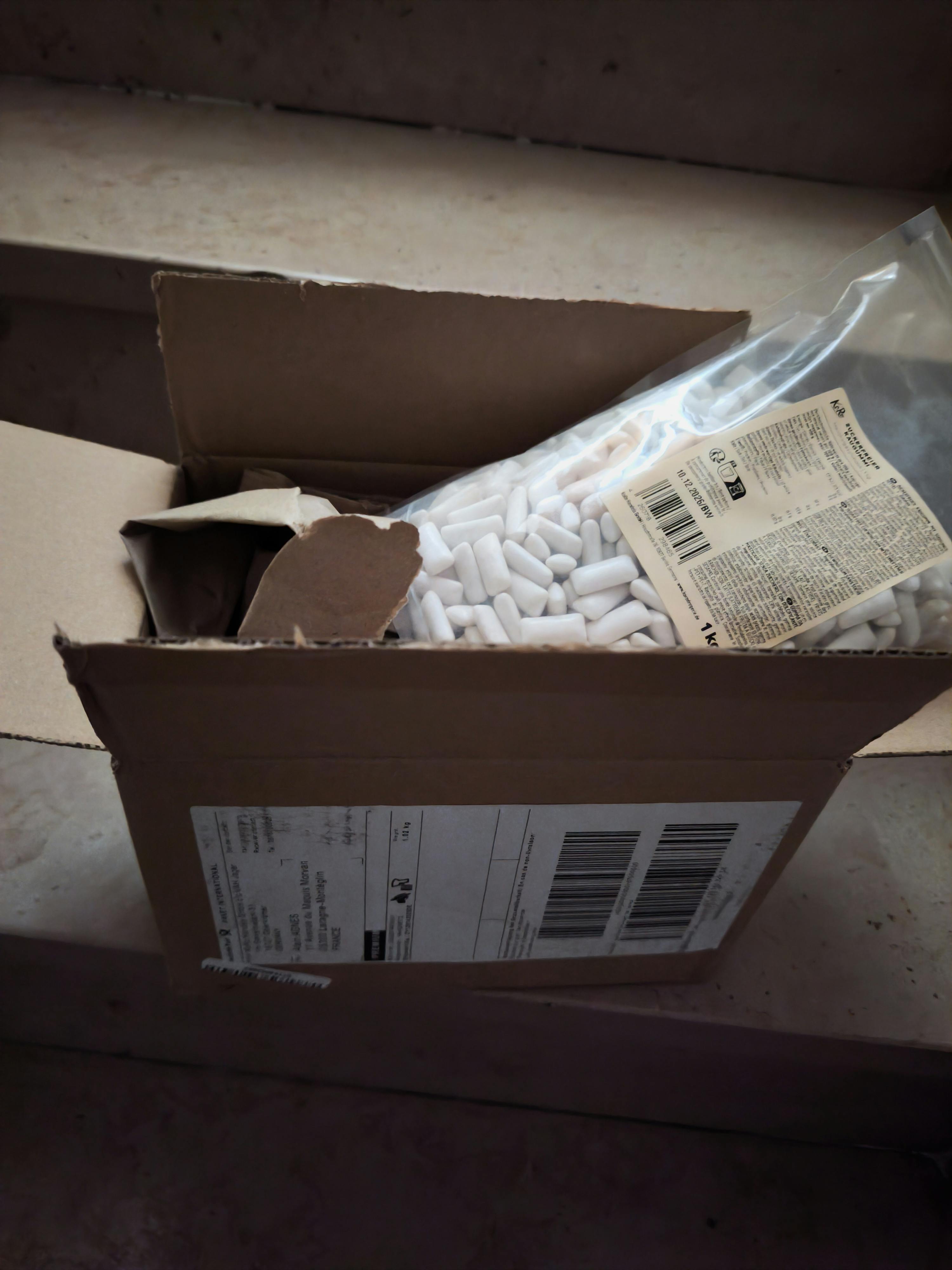 una scatola di cartone aperta contiene un sacchetto di plastica trasparente pieno di piccoli oggetti bianchi a forma di capsula, con etichette visibili sul sacchetto e sulla scatola.