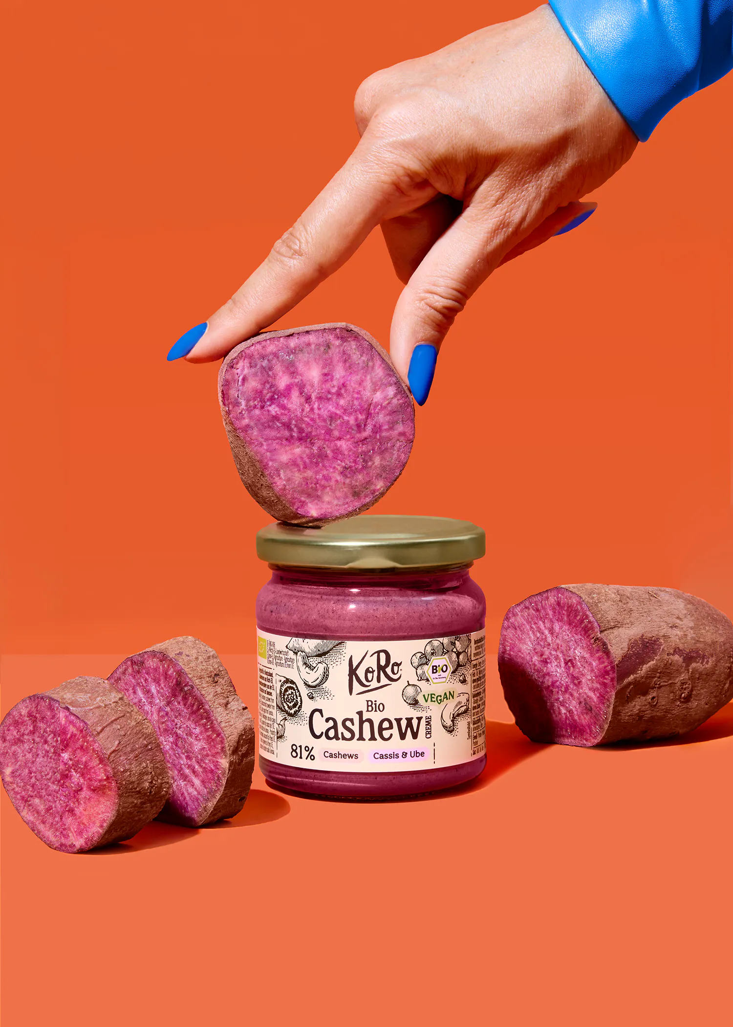 eine hand mit blauen nägeln hält eine scheibe lila süßkartoffel über einem glas lila cashew-creme, umgeben von weiteren süßkartoffelstücken auf orangefarbenem hintergrund.