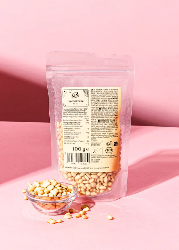 eine Tüte Pinienkerne steht auf einem rosa Tisch neben einer Schüssel Pinienkerne