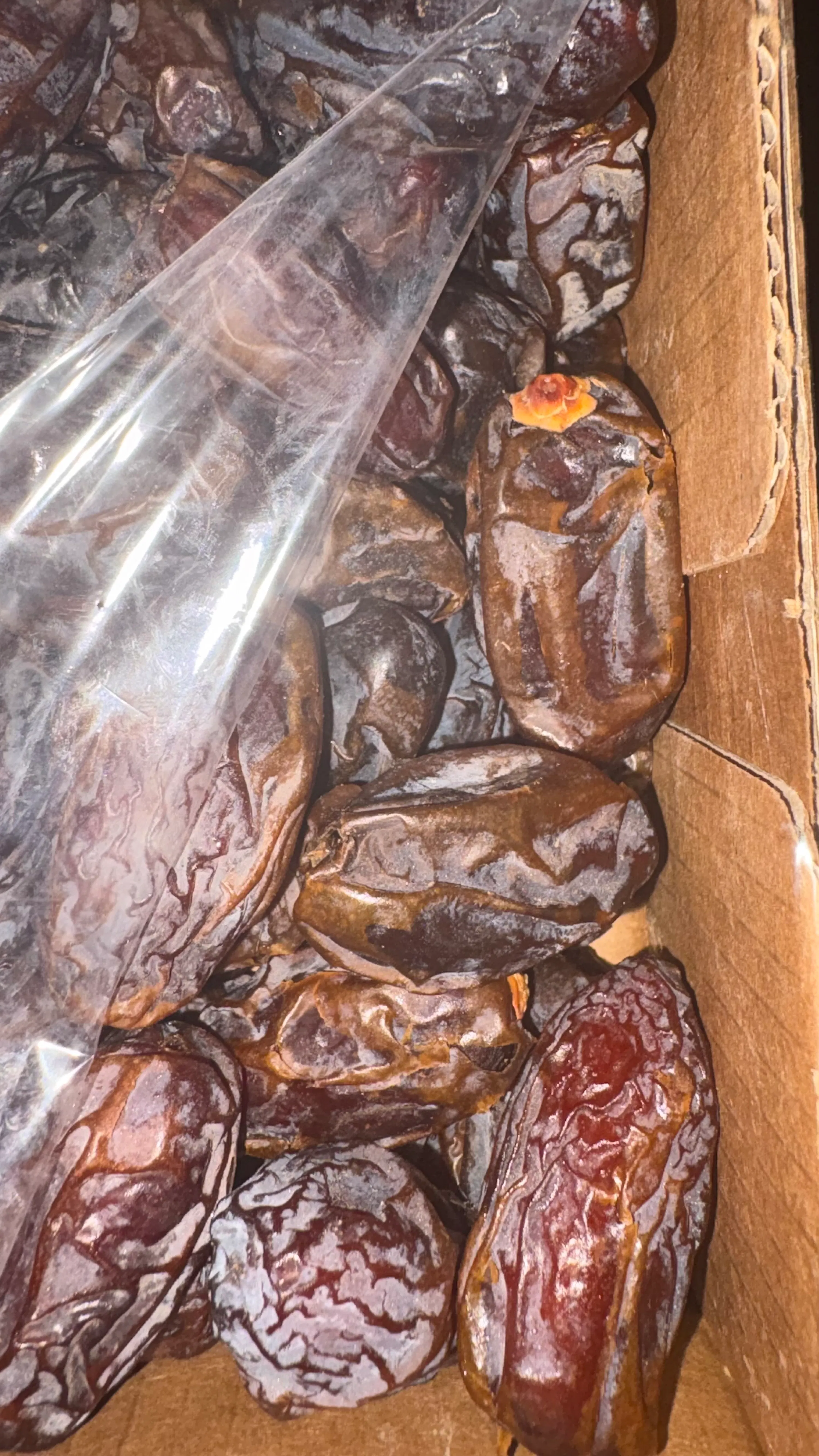 a close-up of many dark brown, wrinkled dates in a cardboard box, with a clear plastic bag partially covering them.
