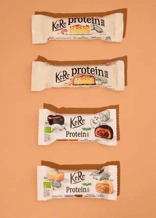 quattro barrette proteiche koro disposte verticalmente su uno sfondo color sabbia, ognuna con il proprio incarto distintivo che mostra il gusto e gli ingredienti.