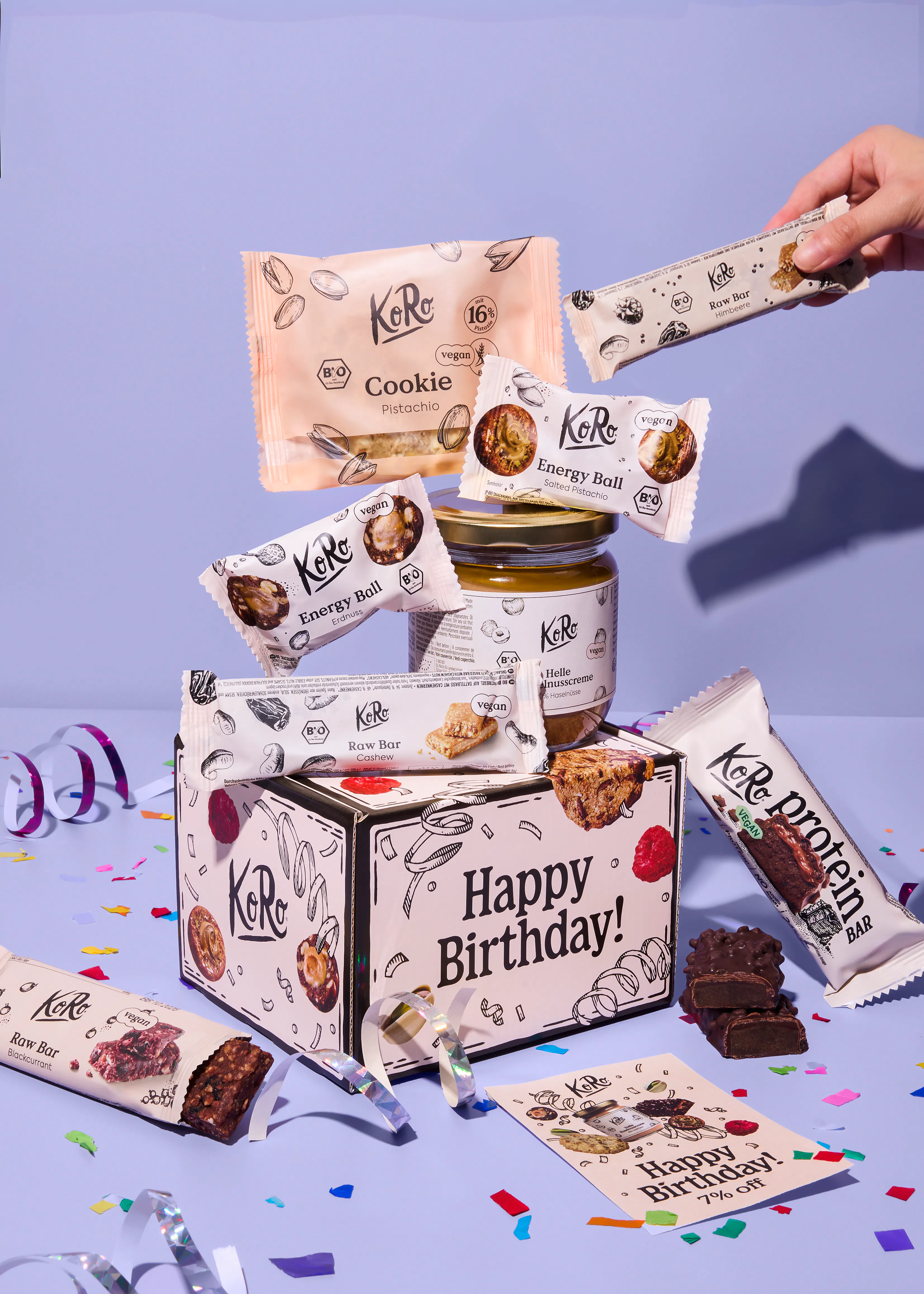 eine geschenkbox von koro mit verschiedenen veganen snacks wie riegeln, energy balls und nusscreme vor einem lila hintergrund mit konfetti.