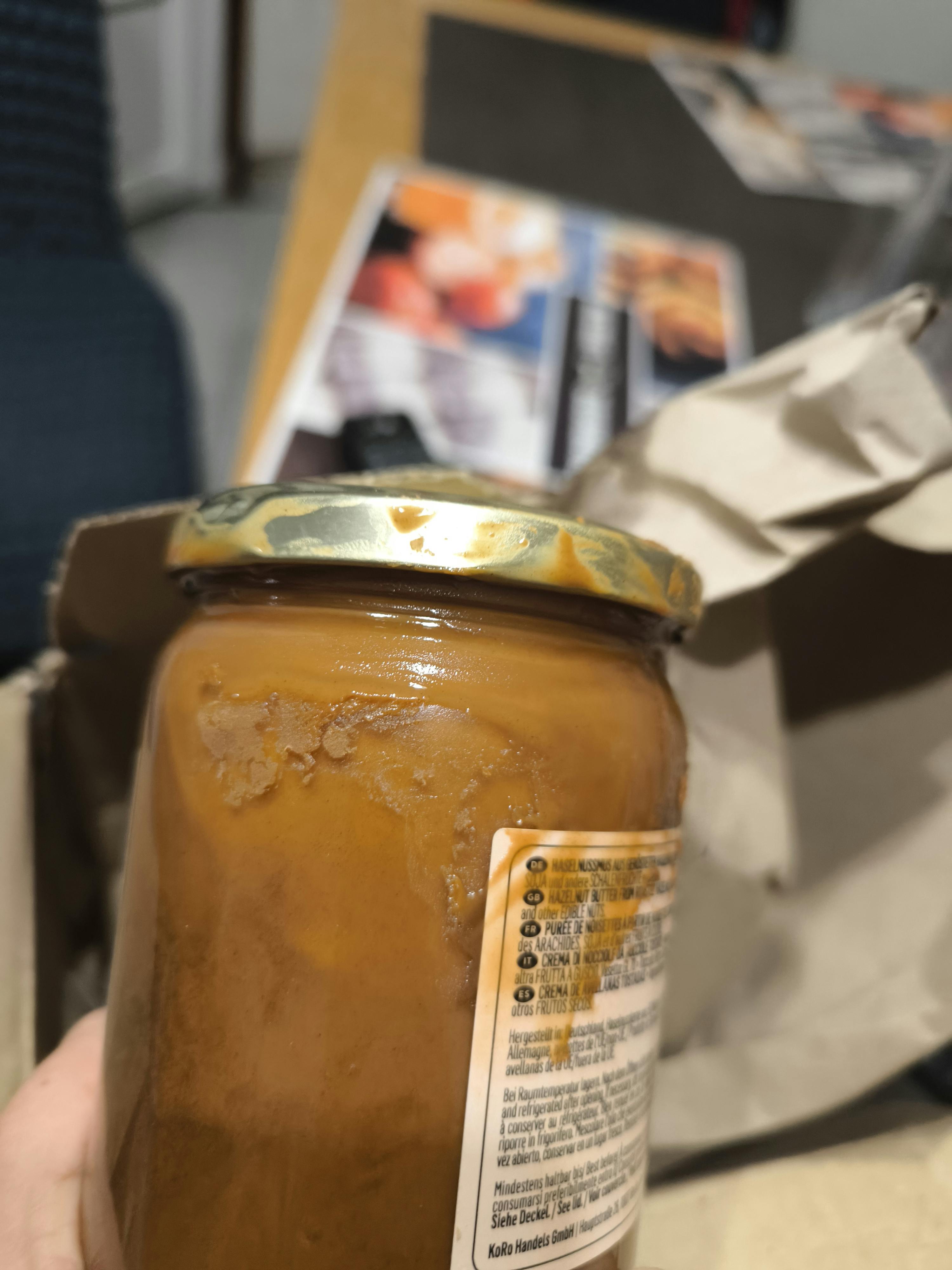 ein glas nussmus mit goldenem deckel und rückständen an der seite, auf dessen etikett mehrsprachige informationen zu sehen sind, steht vor einem unscharfen hintergrund.