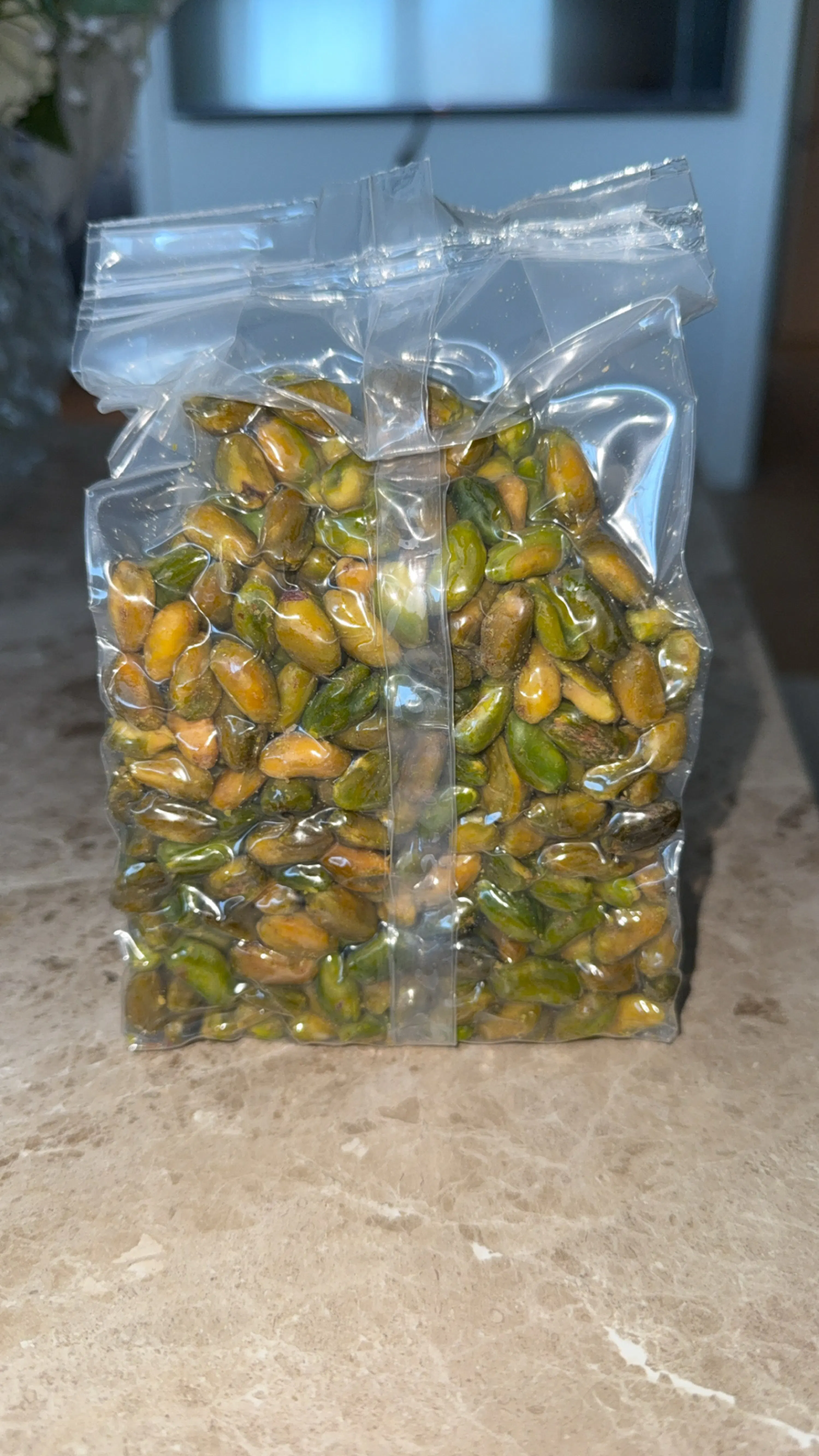 a clear plastic bag filled with shelled green and yellowish-brown pistachios sits on a light brown marble surface.