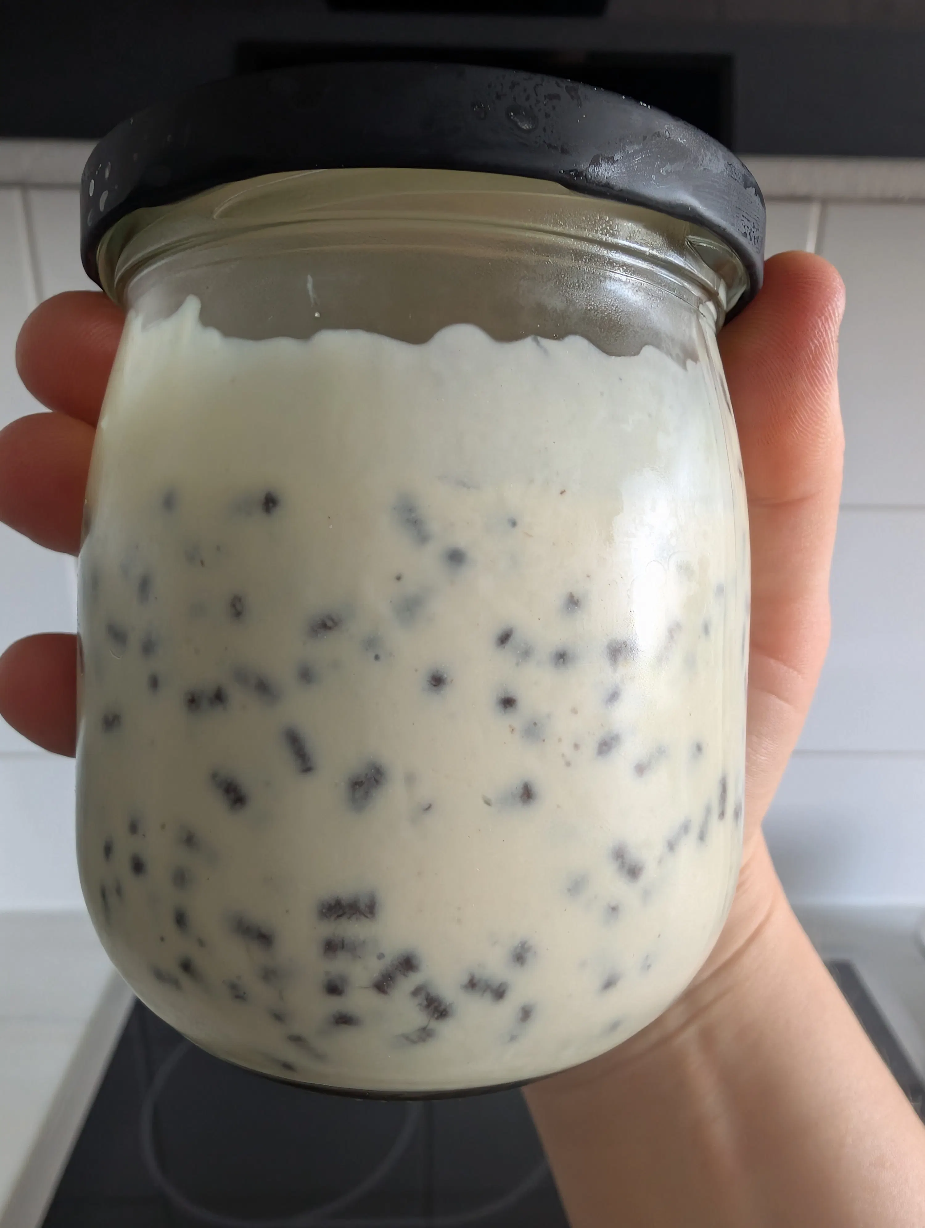 une main tient un pot en verre rempli de yaourt ou de pudding aux graines de chia, avec un couvercle noir.