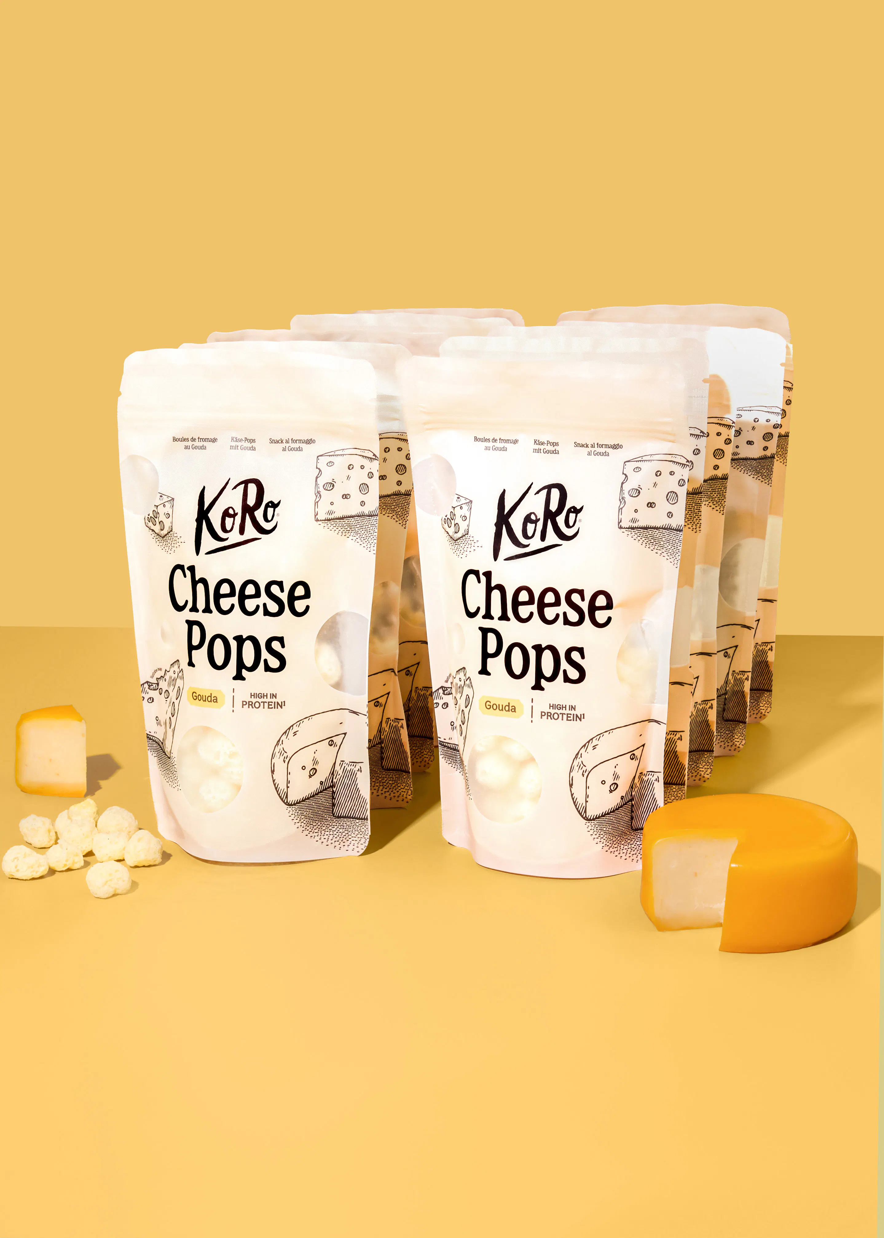 several bags of koro gouda cheese pops arranged on a yellow surface with loose cheese snacks and a small wheel of gouda cheese nearby.
