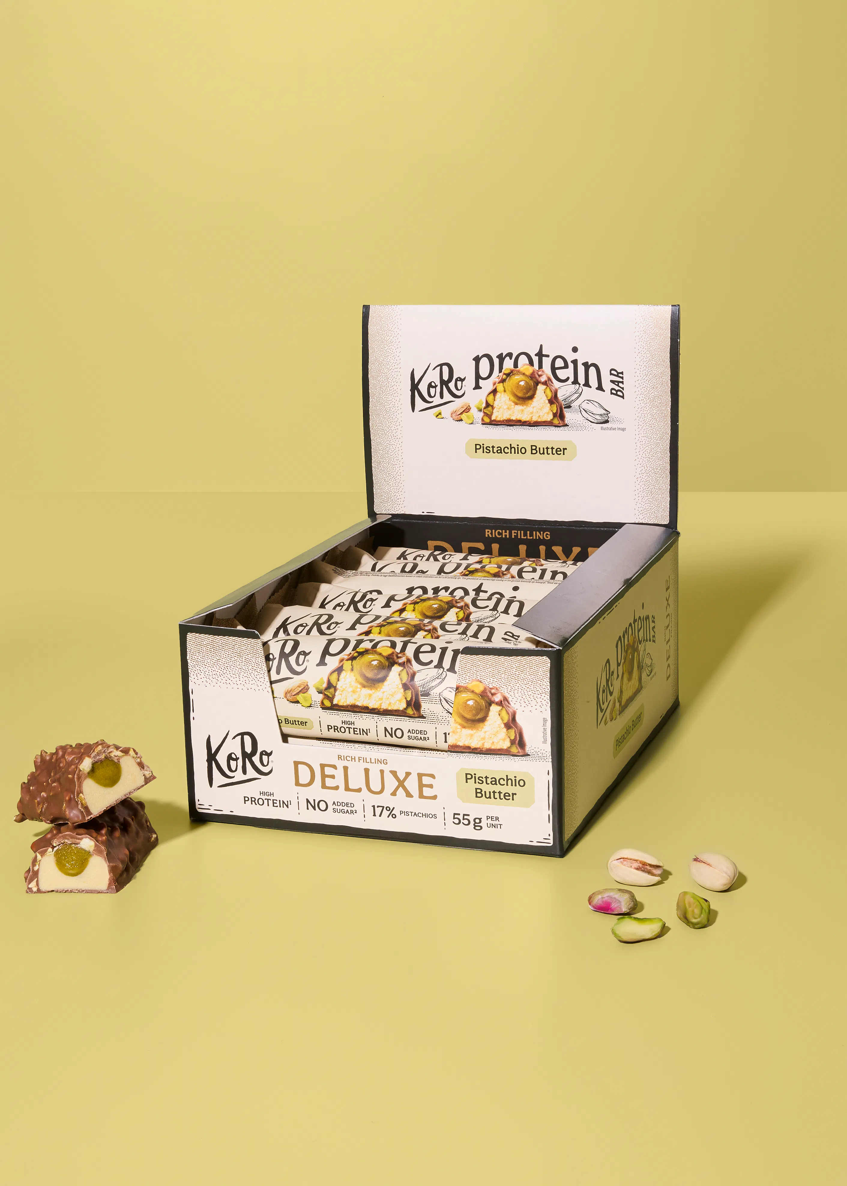 eine offene schachtel koro protein deluxe riegel mit pistazienbutterfüllung, ein halbierter riegel zeigt die grüne füllung, und lose pistazien liegen daneben.