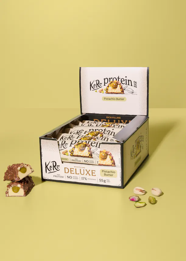eine offene schachtel koro protein deluxe riegel mit pistazienbutterfüllung, ein halbierter riegel zeigt die grüne füllung, und lose pistazien liegen daneben.