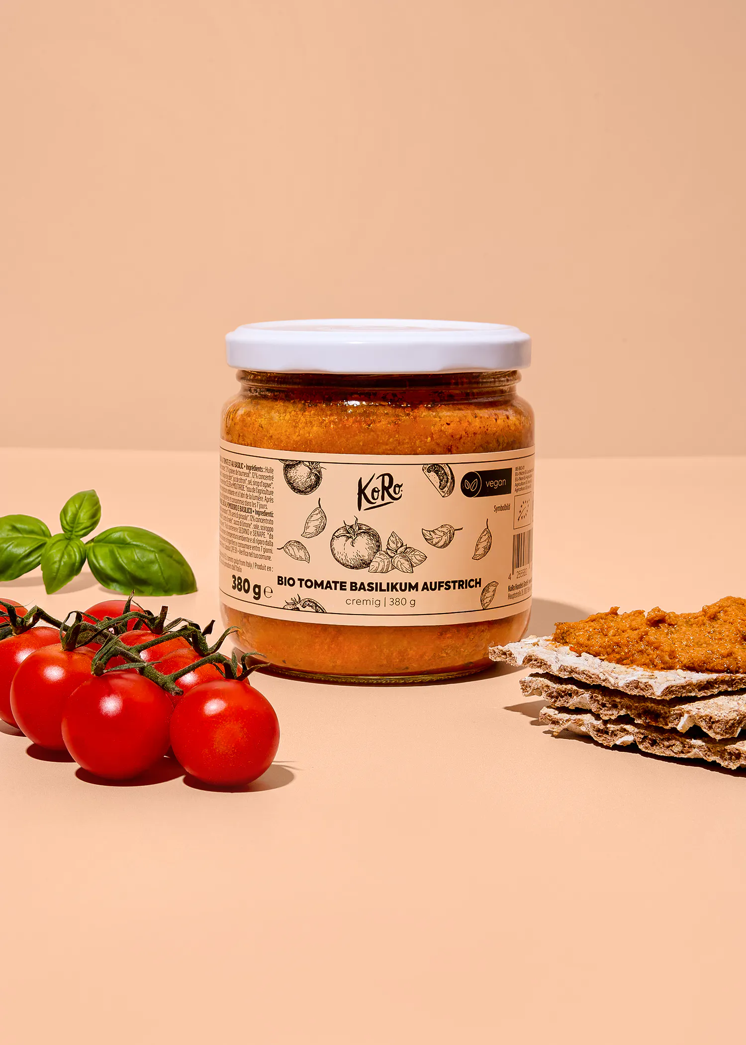 un pot de tartinade bio tomate basilic koro posé sur un fond beige avec des tomates cerises fraîches, des feuilles de basilic et des craquelins tartinés à côté.