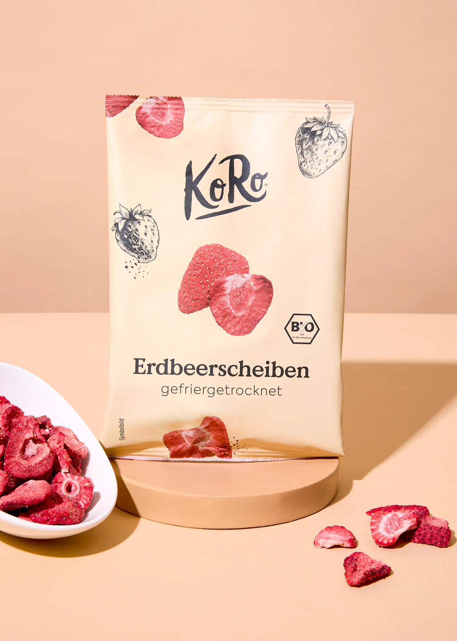 eine packung koro gefriergetrocknete erdbeerscheiben steht auf einem podest, daneben eine schale mit weiteren erdbeerscheiben und einige verstreute stücke auf beigem hintergrund.
