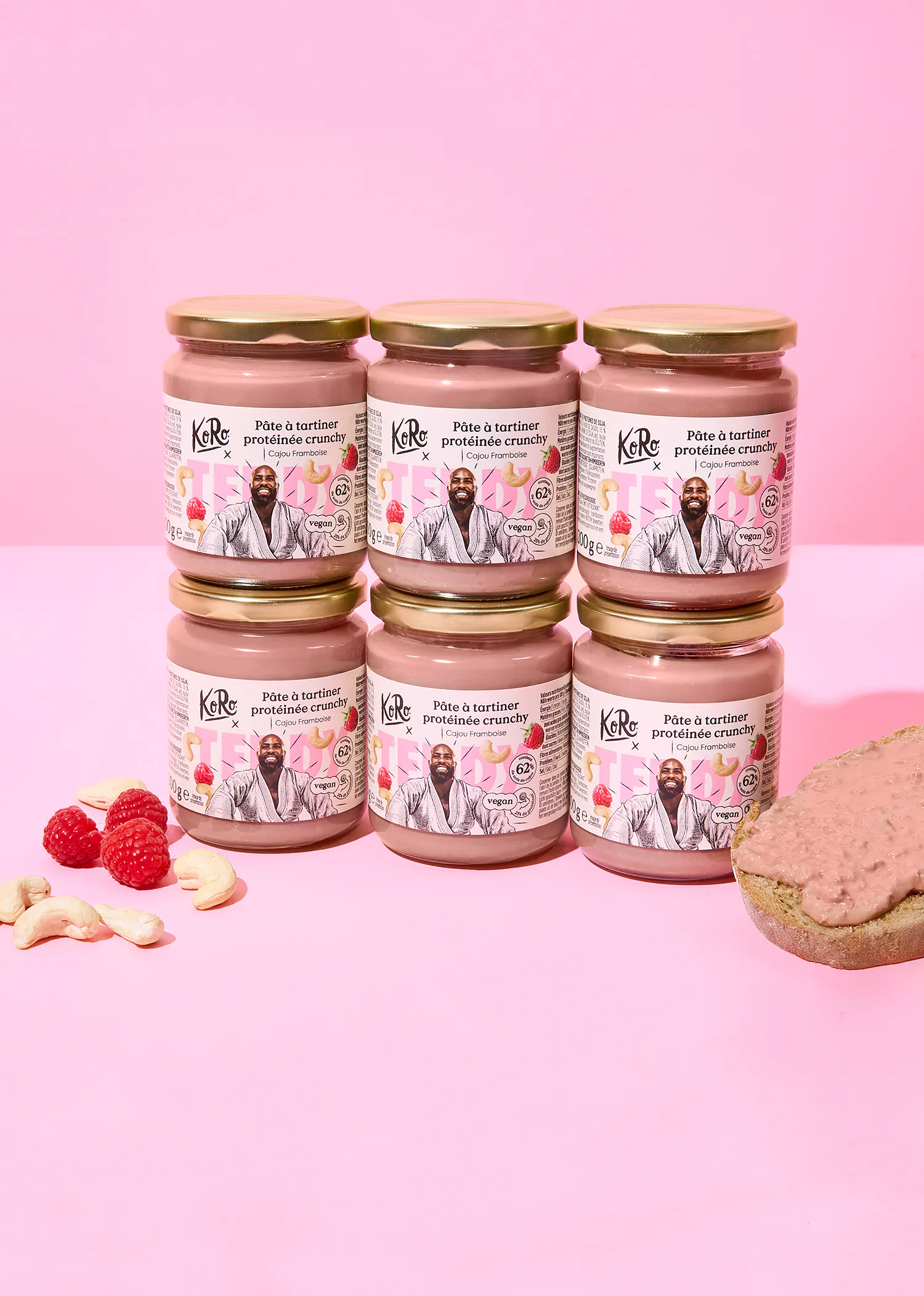 sechs gläser rosa koro knuspriger proteinaufstrich mit cashew und himbeere, umgeben von frischen himbeeren, cashewnüssen und einer scheibe brot mit dem aufstrich.