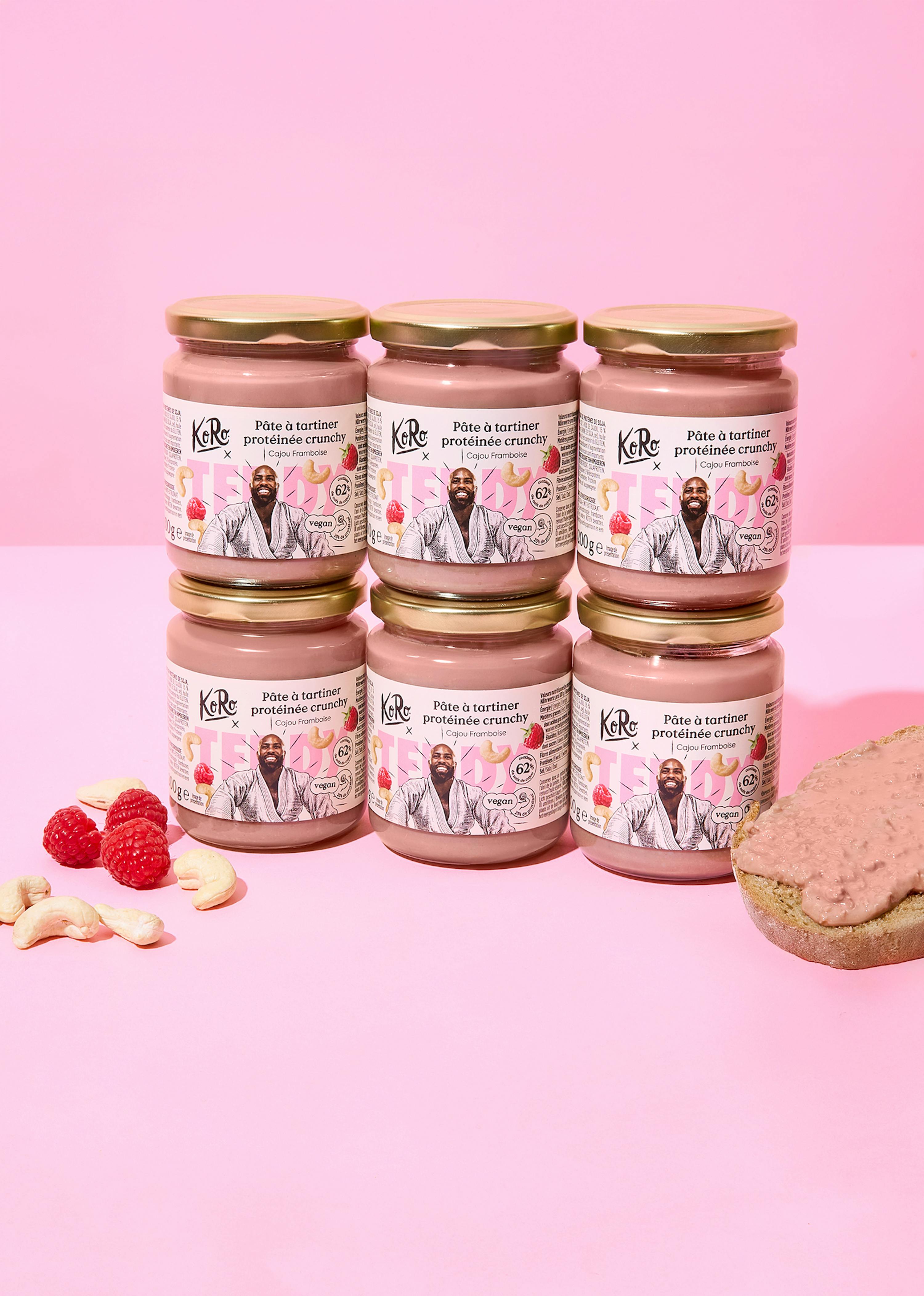 six pots de pâte à tartiner protéinée crunchy cajou framboise koro sont disposés sur un fond rose, accompagnés de framboises, de noix de cajou et d'une tranche de pain tartinée.