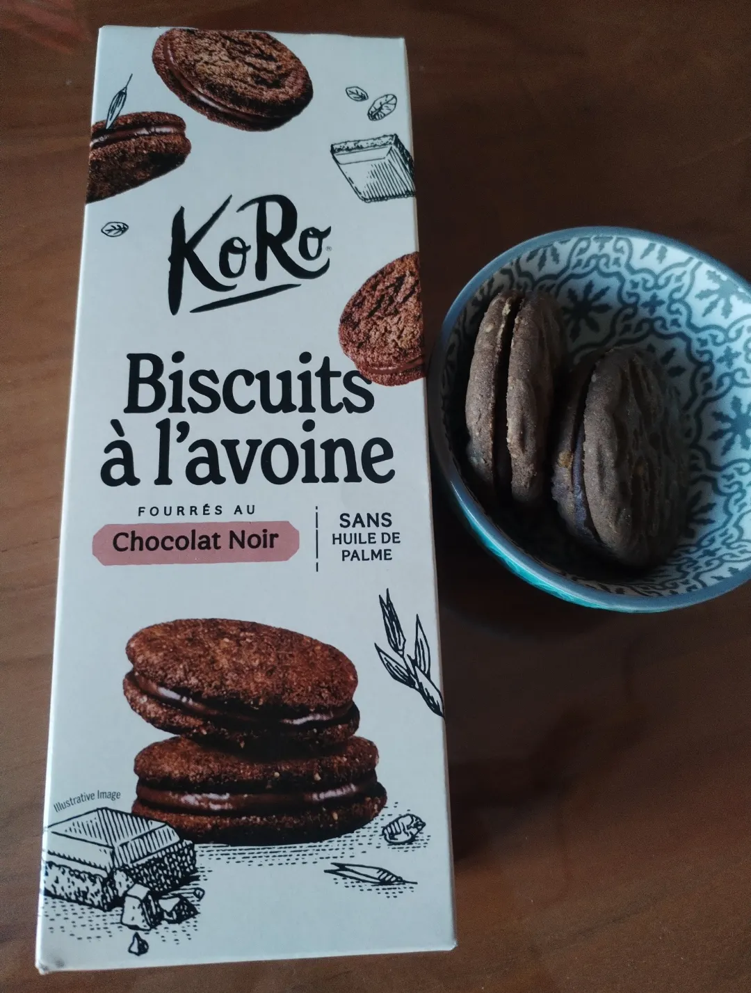 eine verpackung von koro haferkeksen mit zartbitterschokoladenfüllung steht neben einer kleinen schale mit zwei der kekse auf einem holztisch.