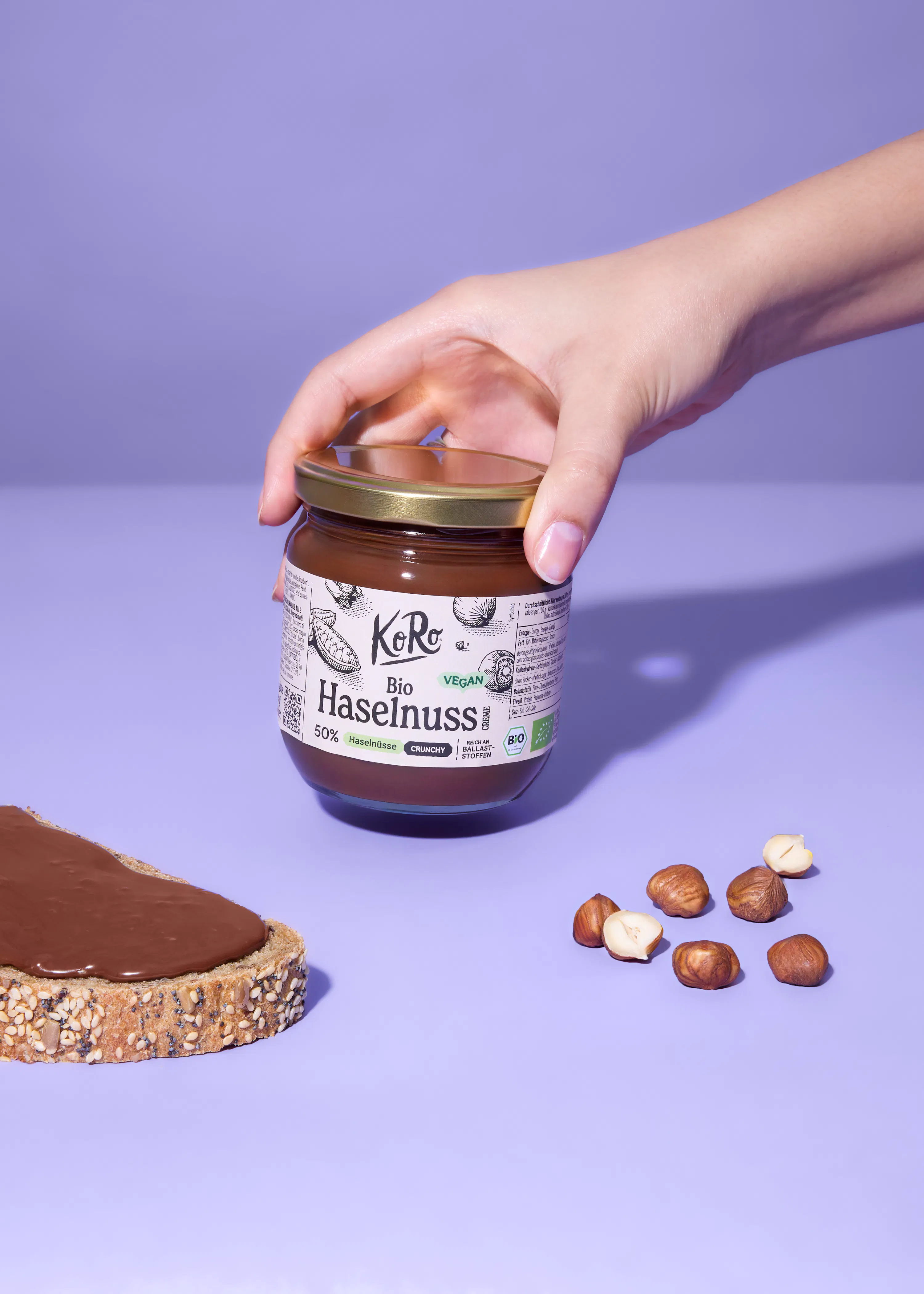 eine hand hält ein glas koro bio haselnuss-brotaufstrich, daneben liegen ein brot mit dem aufstrich und ganze sowie halbierte haselnüsse auf einem lila hintergrund.