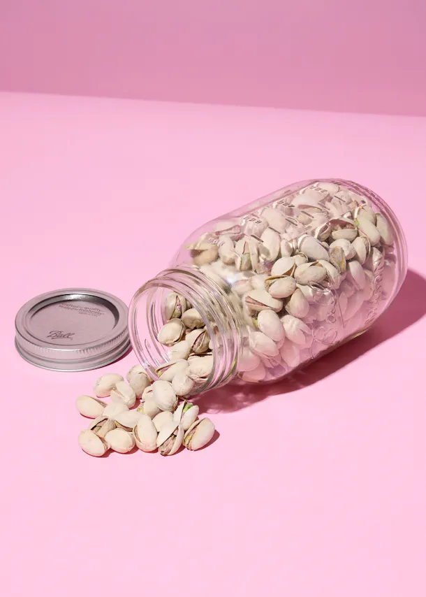 a glass jar of pistachios in their shells lies on its side on a pink background, with some pistachios spilled out and the silver lid next to it.
