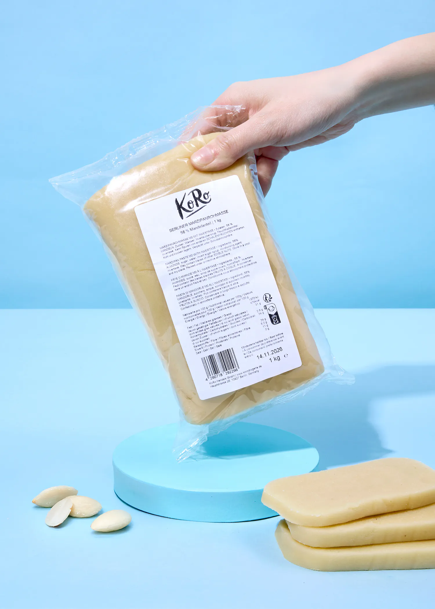 eine hand hält eine durchsichtige plastikverpackung mit berliner marzipanrohmasse über einem hellblauen ständer, auf dem auch lose marzipanstücke und blanchierte mandeln liegen.