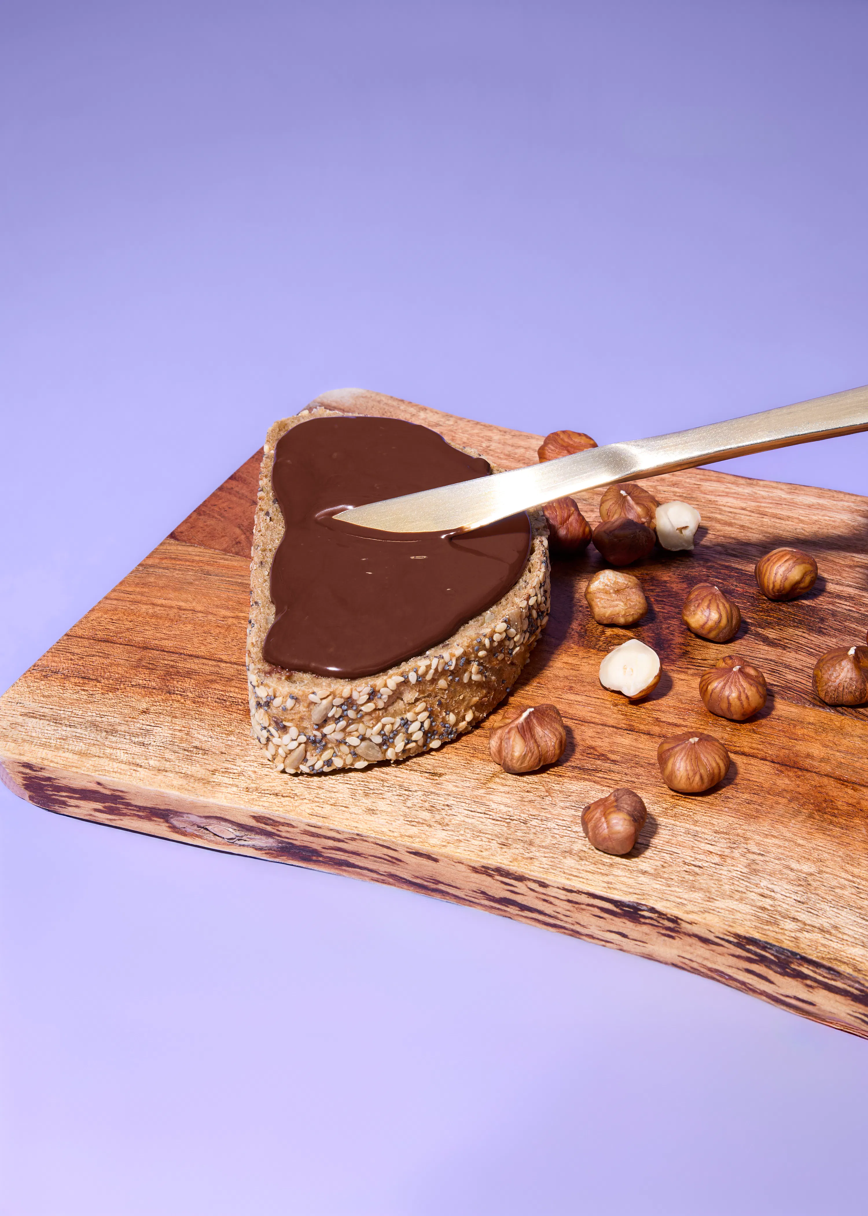 eine scheibe brot mit schokoladenaufstrich und einem goldenen messer auf einem holzbrett mit haselnüssen.