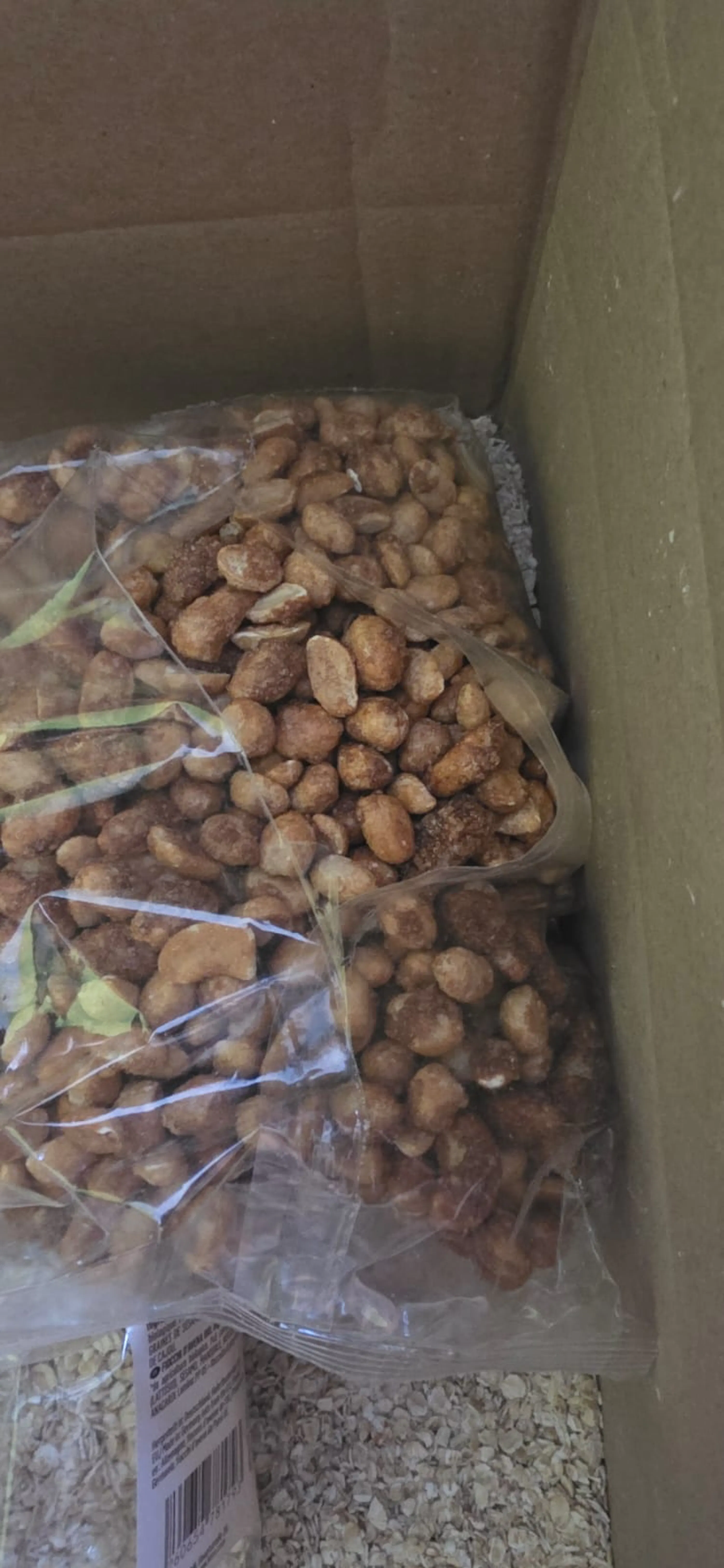 deux sacs en plastique transparents remplis de cacahuètes grillées ou caramélisées sont visibles dans une boîte en carton, avec une étiquette et des flocons d'avoine en dessous.