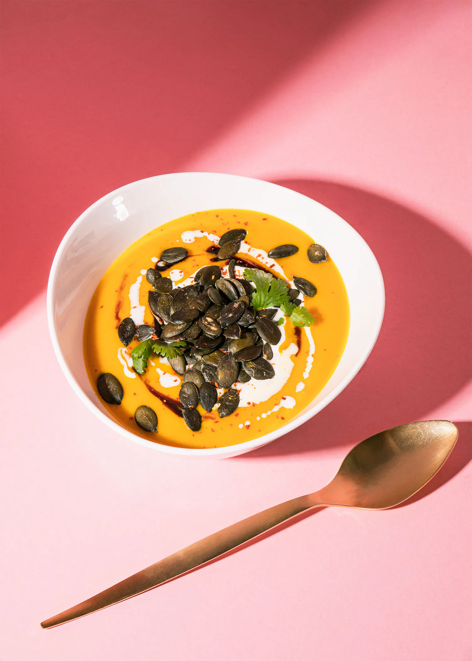 un bol de soupe avec des graines de citrouille et une cuillère sur une table rose