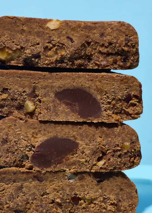 une pile de quatre barres protéinées ou de collation brunes, texturées, avec des morceaux de noix et de gros morceaux de chocolat, sur un fond bleu clair.