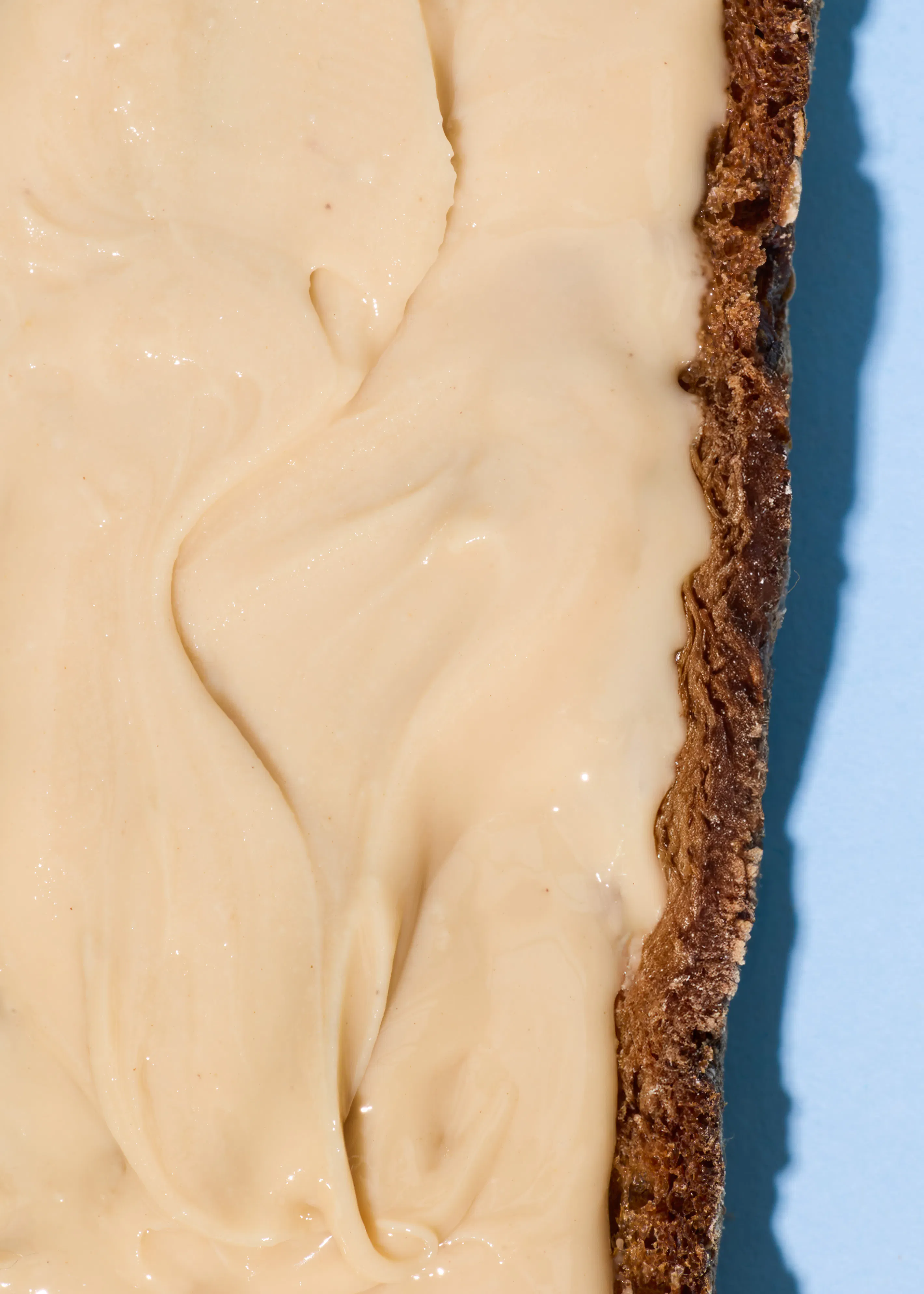 eine nahaufnahme einer hellen, cremigen masse mit sanften wirbeln, die einen dunklen, strukturierten rand bedeckt, vor einem hellblauen hintergrund.