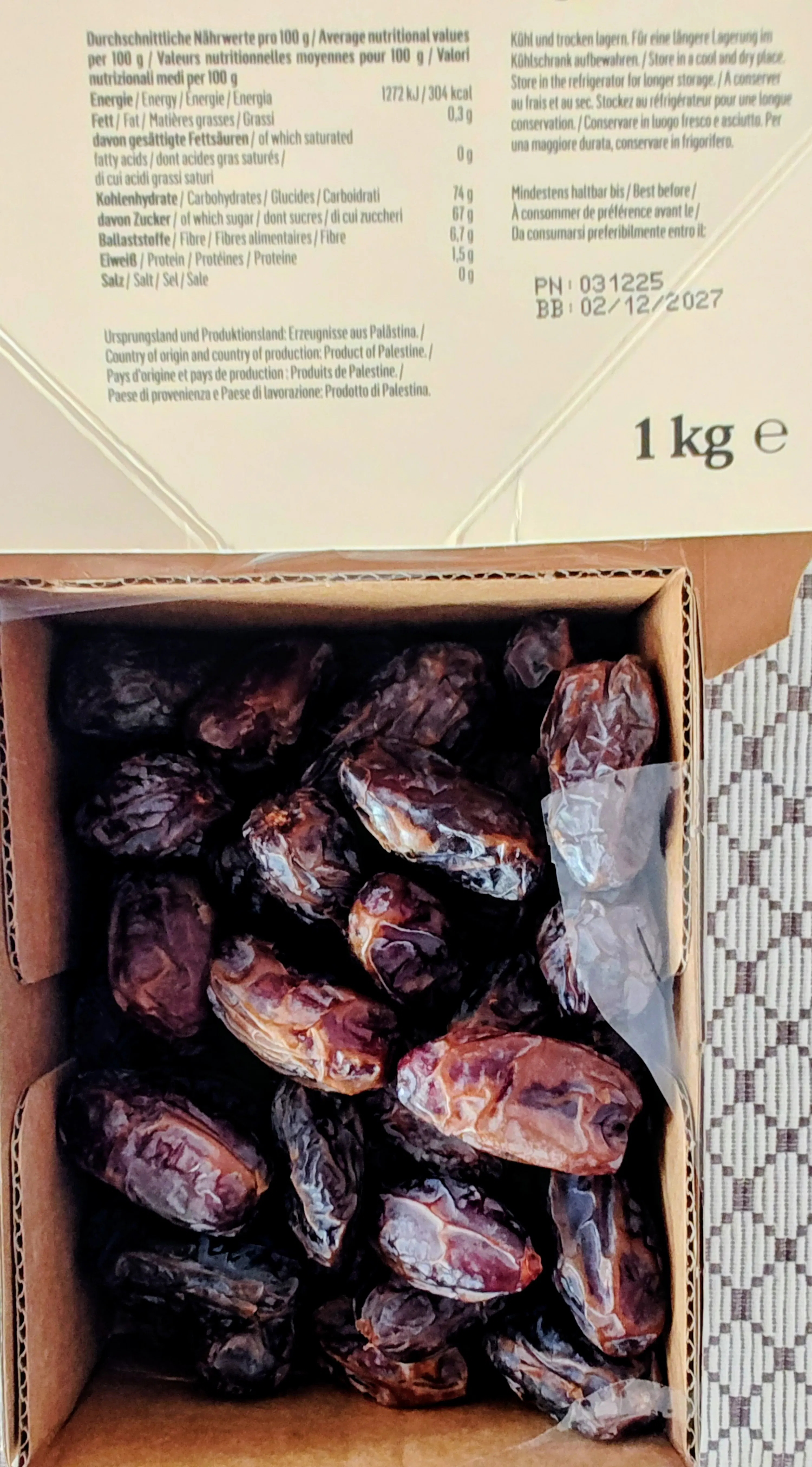 eine offene pappschachtel, gefüllt mit vielen dunklen, runzligen datteln, wobei ein teil des produktetiketts mit nährwertangaben, dem herkunftsland palästina und dem gewicht von 1 kg sichtbar ist.