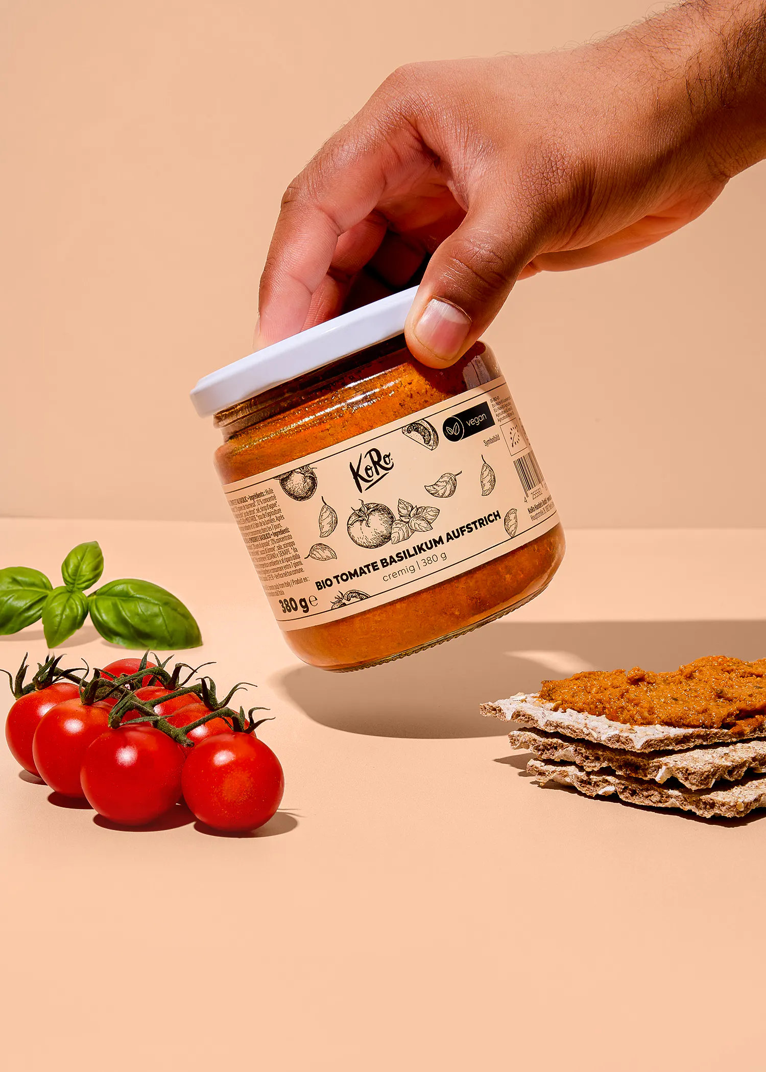 eine hand hält ein glas koro bio tomate basilikum aufstrich neben frischen kirschtomaten und einem mit aufstrich bestrichenen knäckebrot auf beigem hintergrund.