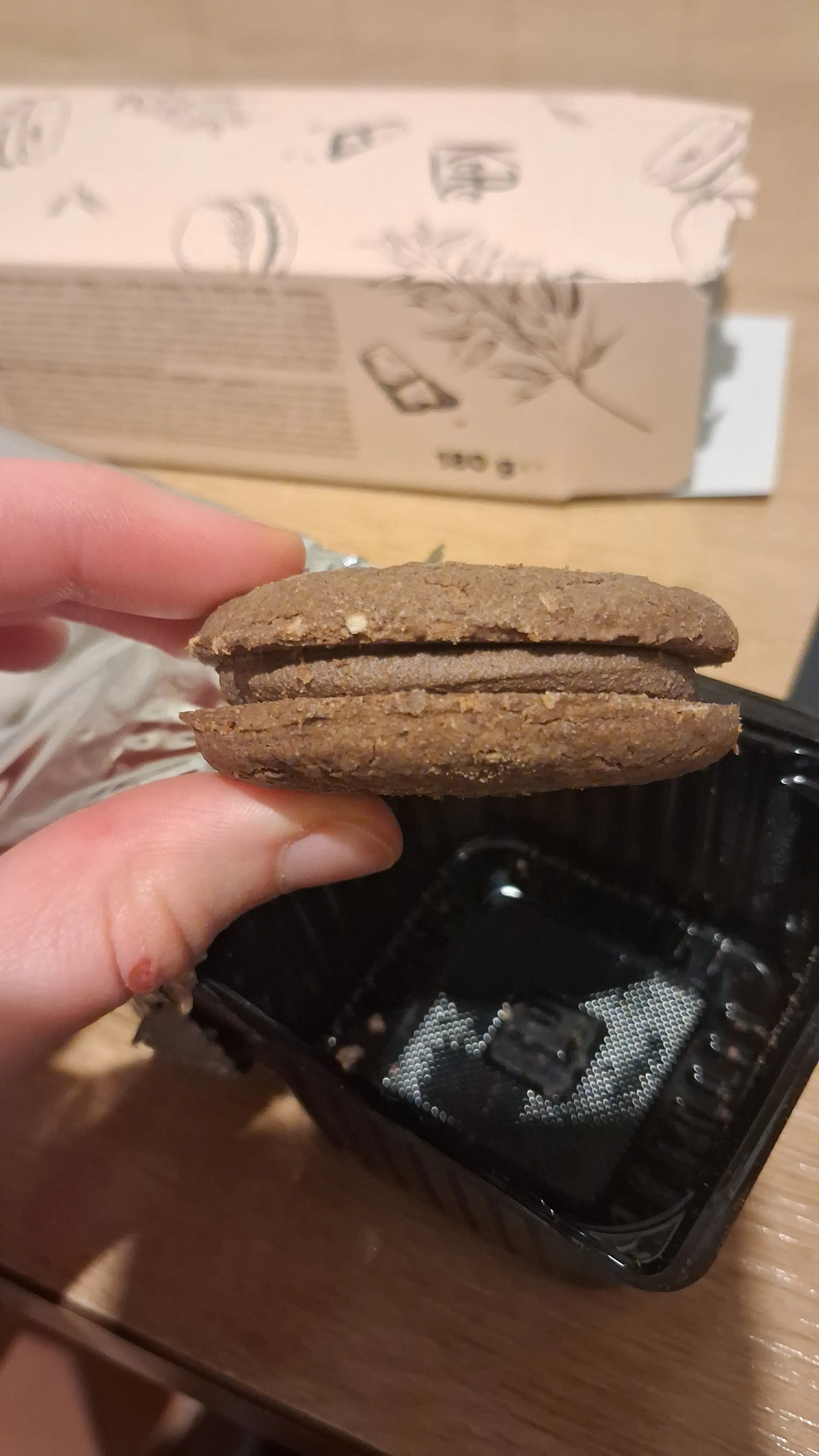 une main tient un biscuit sandwich au chocolat avec une garniture crémeuse, devant un emballage en carton et un récipient en plastique noir.