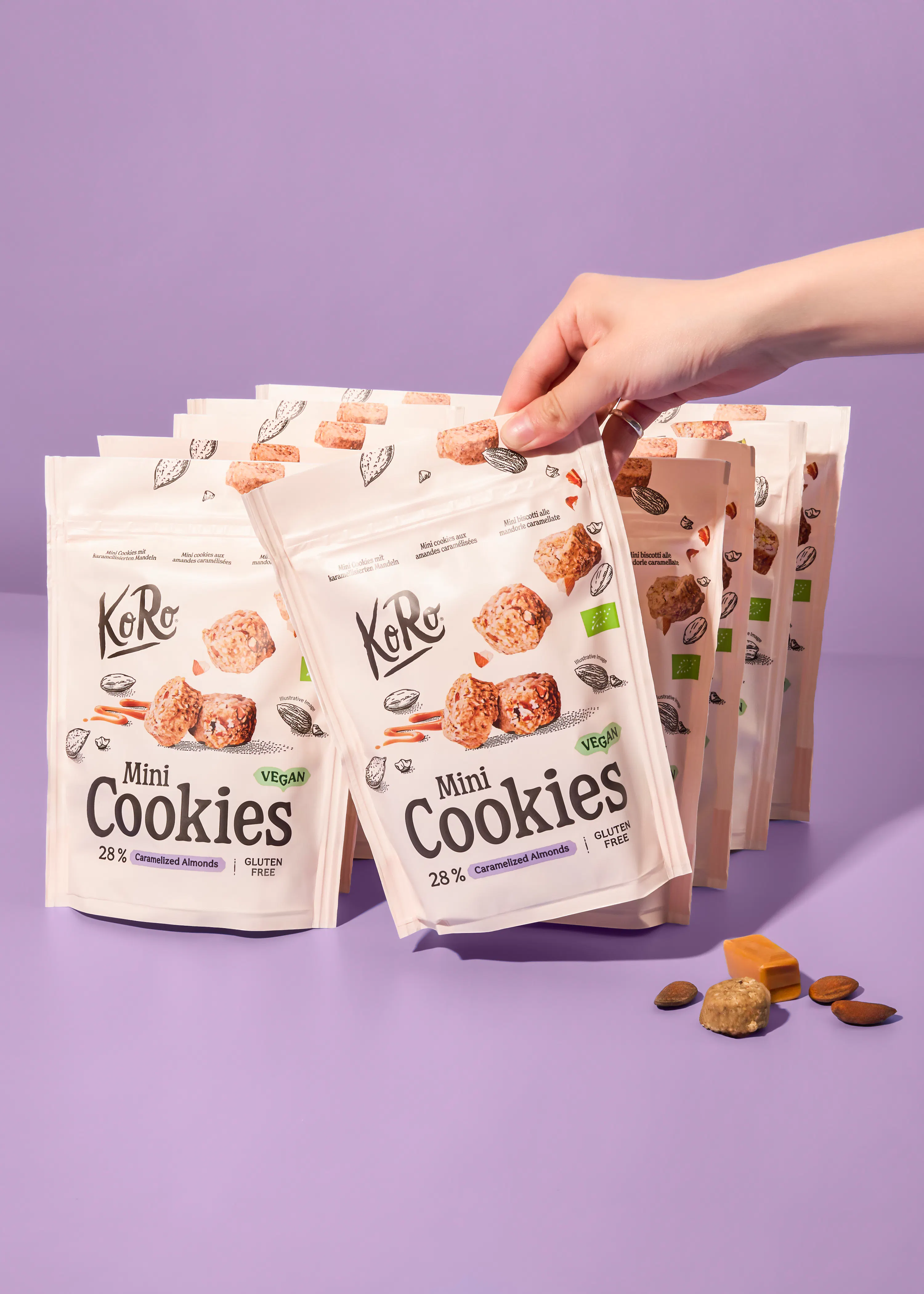 eine hand greift nach einer weißen verpackung von koro mini cookies mit karamellisierten mandeln, umgeben von weiteren packungen und einzelnen mandeln und karamellstücken auf einem lila hintergrund.