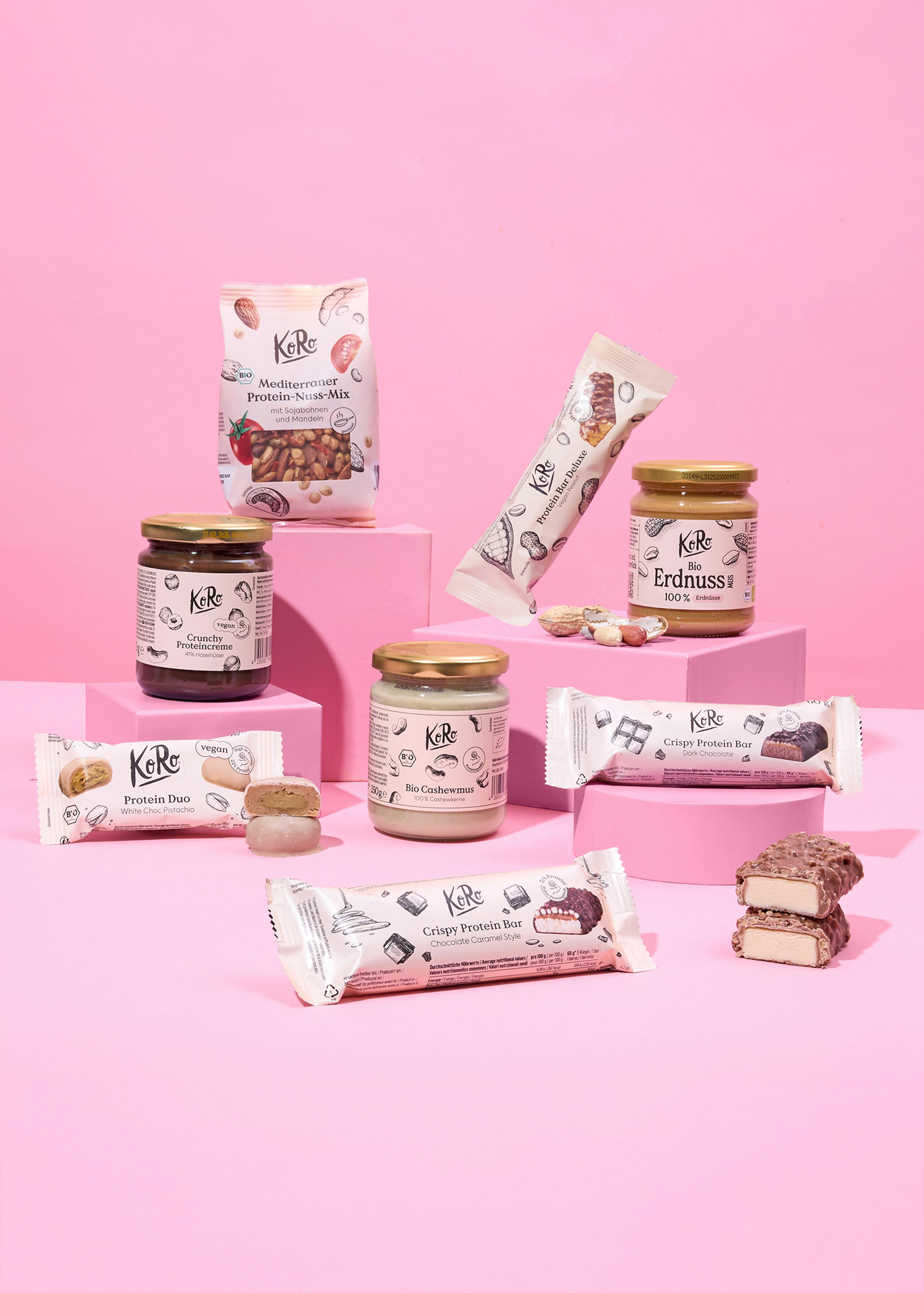 une variété de produits protéinés koro, y compris des barres, des mélanges de noix et des pâtes à tartiner, sont présentés sur des socles roses sur un fond rose uni.