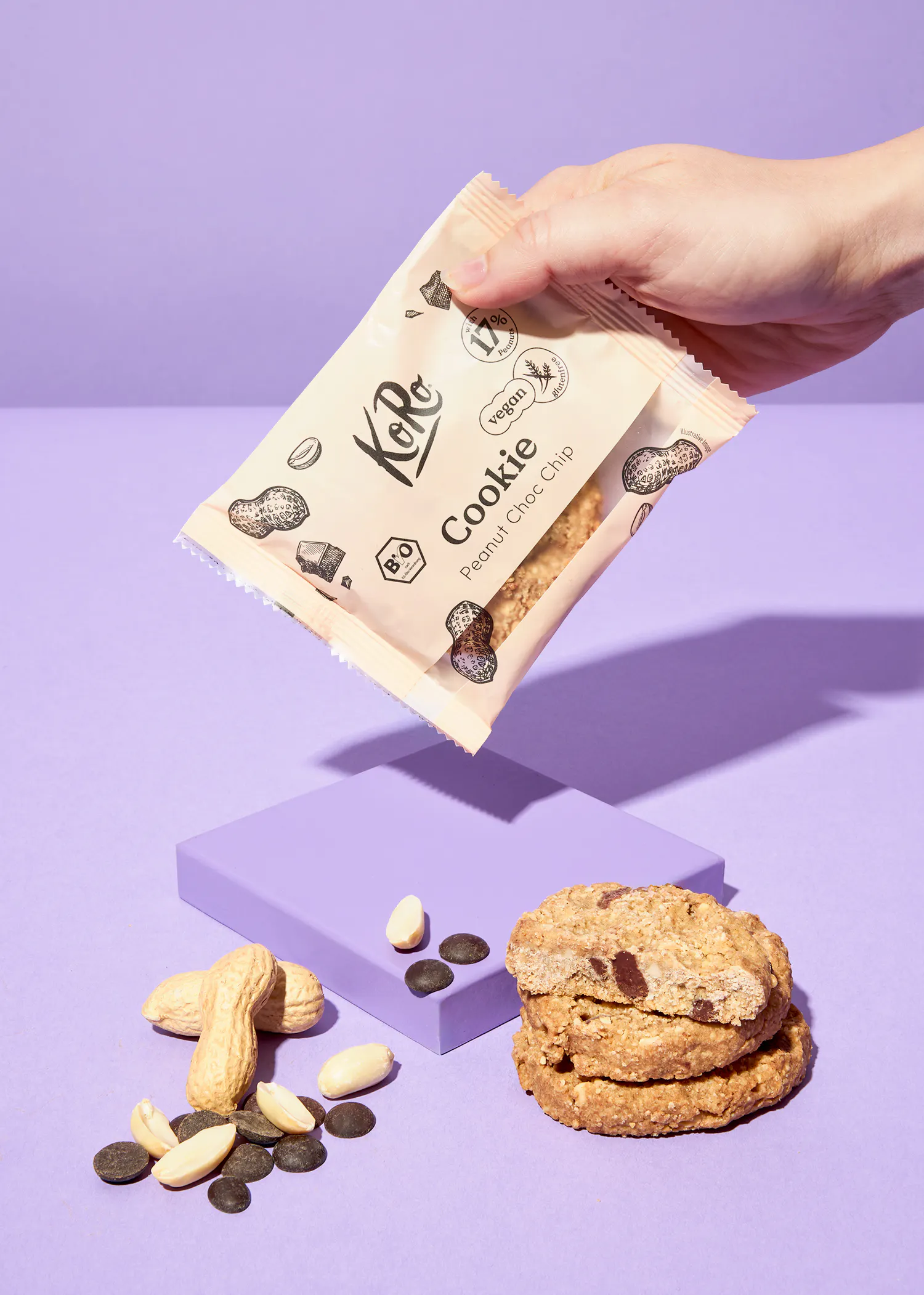 une main tient un paquet de biscuits koro cookie peanut choc chip végétaliens et sans gluten, tandis que des cacahuètes, des pépites de chocolat et une pile de biscuits sont disposés sur un fond violet.