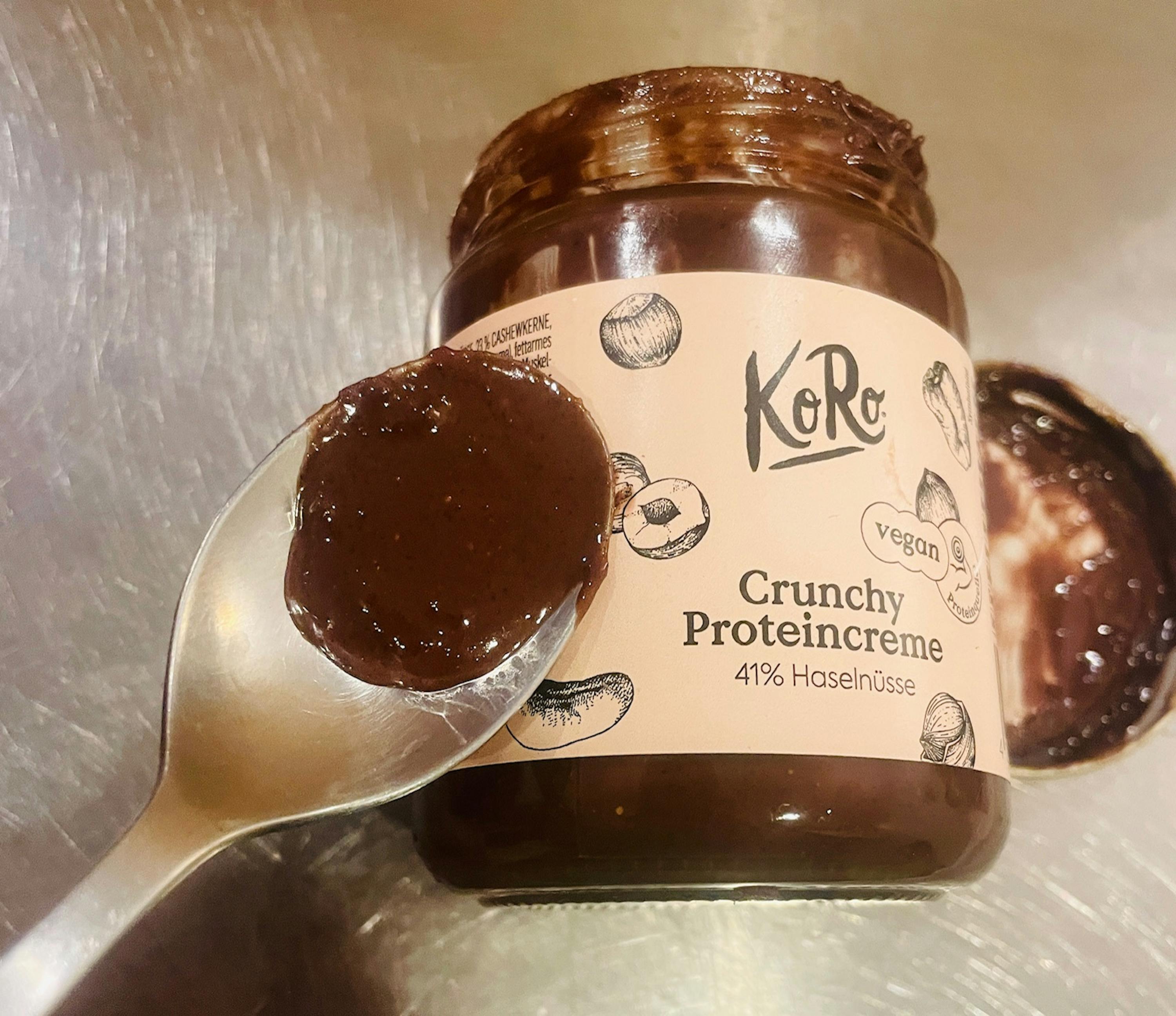un pot de pâte à tartiner protéinée croquante koro au chocolat et aux noisettes, avec une cuillère remplie de la même pâte à côté, sur une surface métallique.