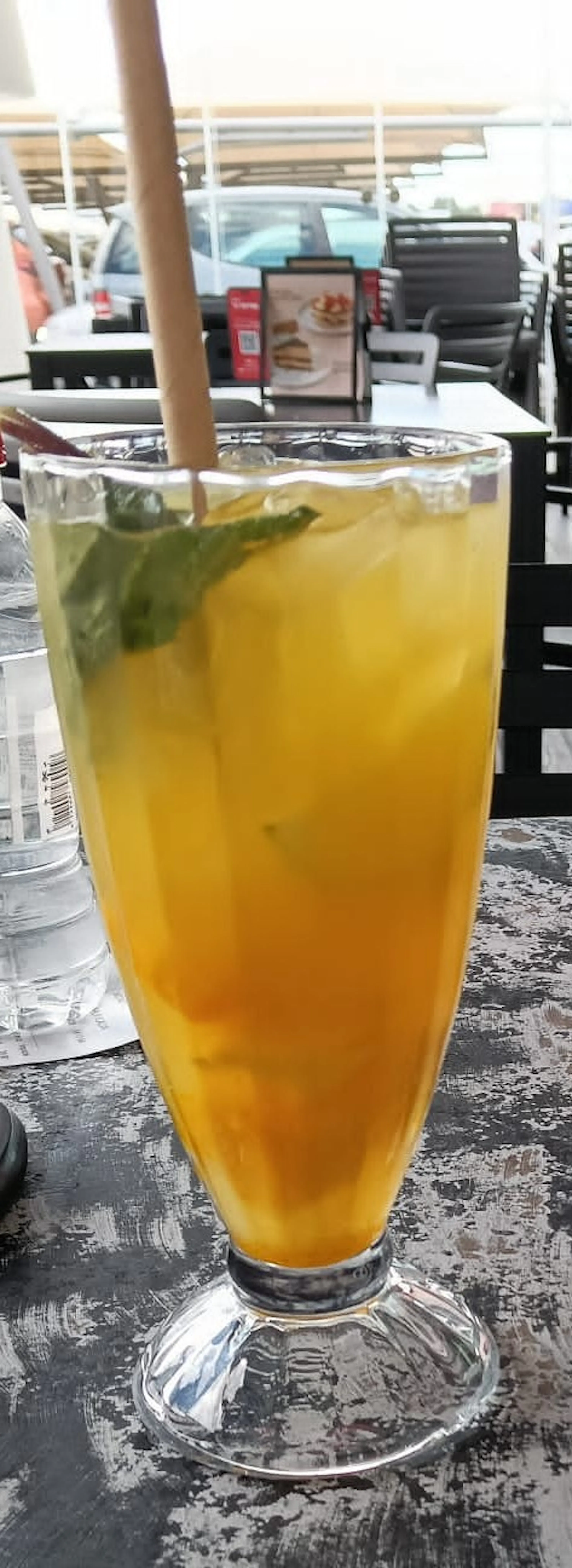 un grand verre de boisson jaune orangé avec des glaçons, des feuilles de menthe et une paille en bois, posé sur une table.