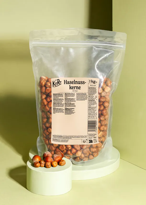 a clear plastic bag of koro 1kg hazelnut kernels sits on a light green stand, with a few whole and shelled hazelnuts on a smaller stand in the foreground.