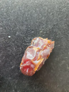 eine einzelne, faltige, rötlich-braune dattelfrucht mit feinen fasern liegt auf einer dunklen, körnigen oberfläche.