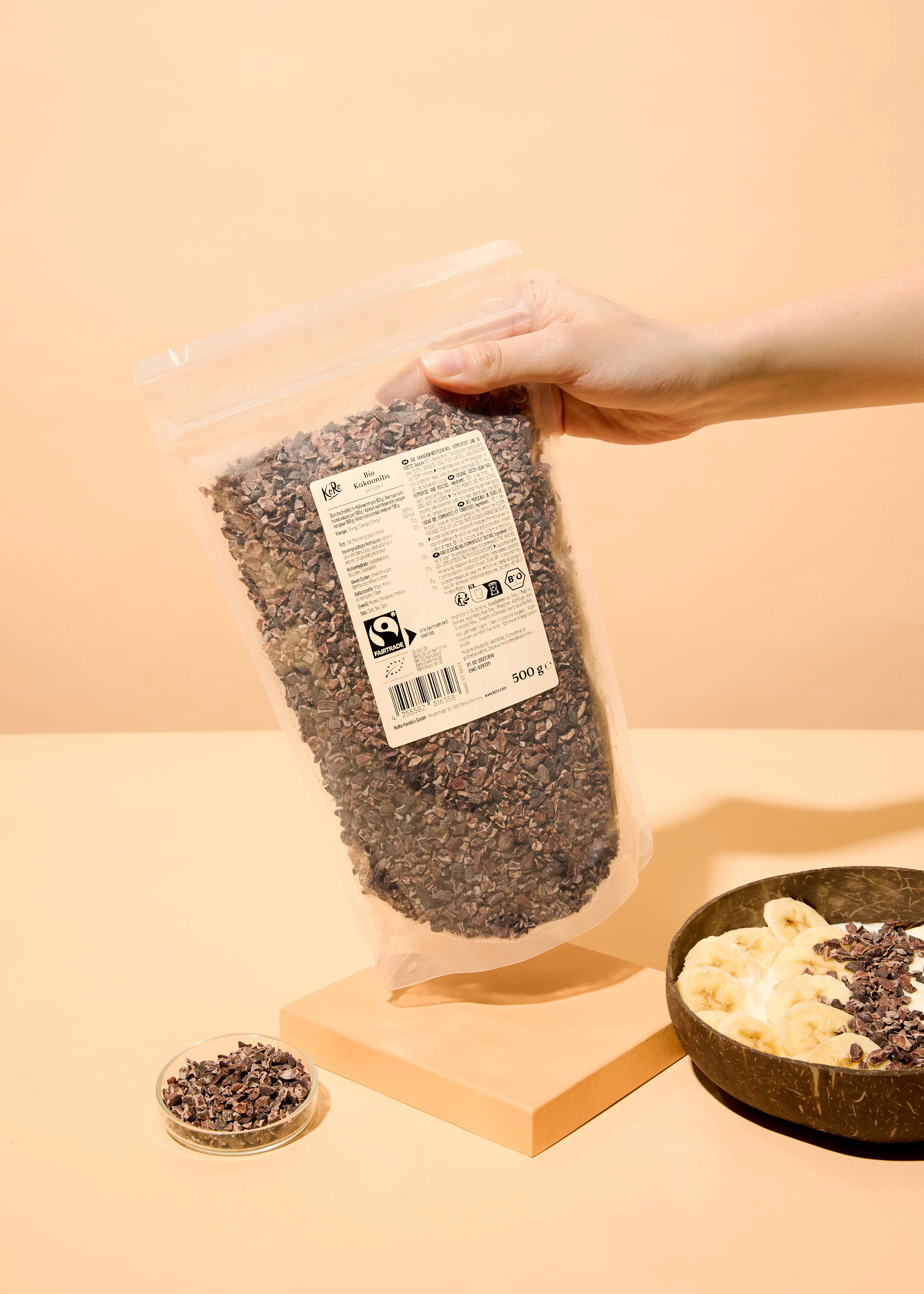 une main tient un sac transparent de fèves de cacao concassées bio, avec un bol de yaourt, bananes et fèves de cacao, et un petit plat de fèves de cacao sur une surface beige.