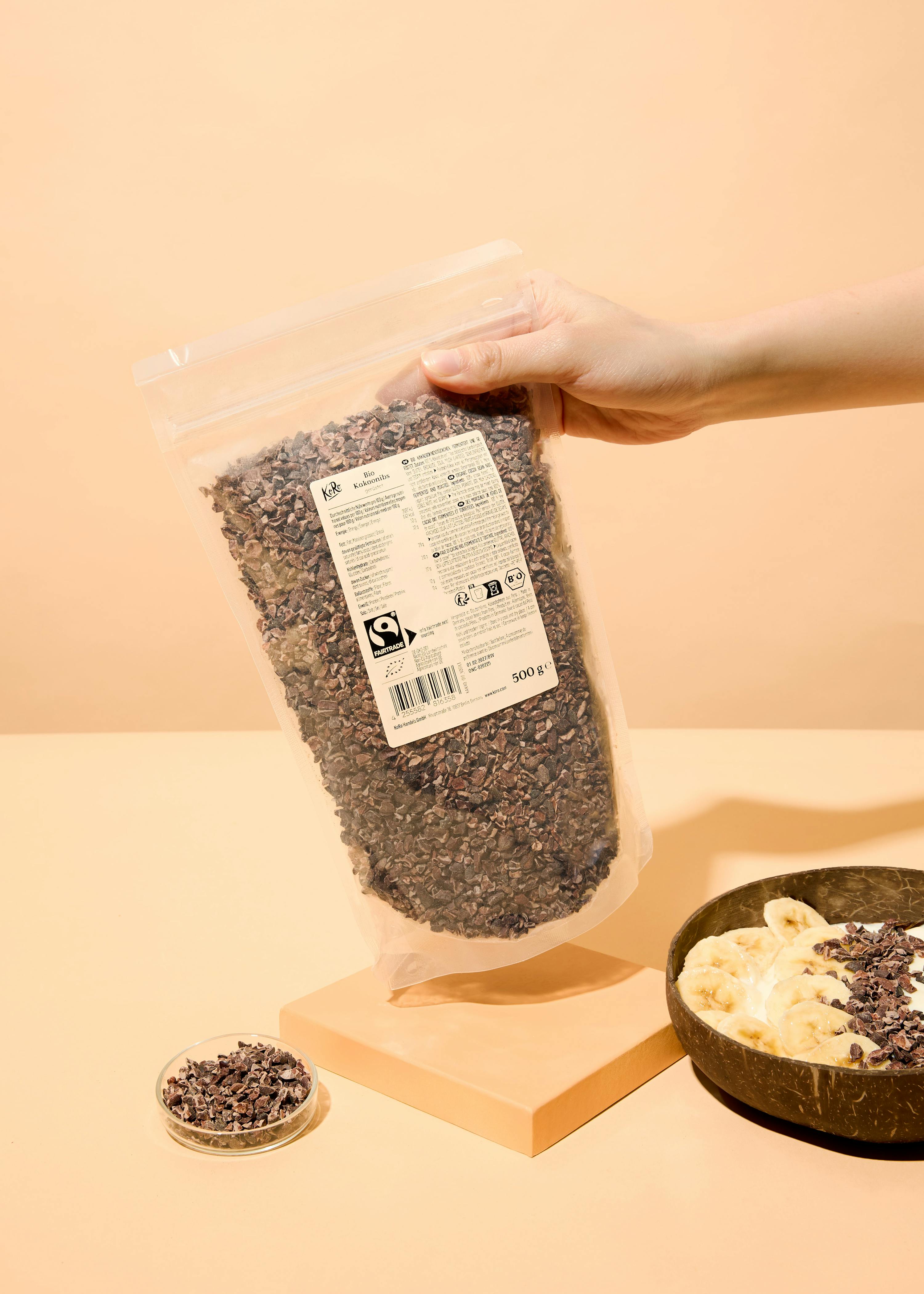 a hand holds a clear bag of cocoa nibs, with a small dish of nibs and a coconut bowl of yogurt, bananas, and nibs on a light orange surface.