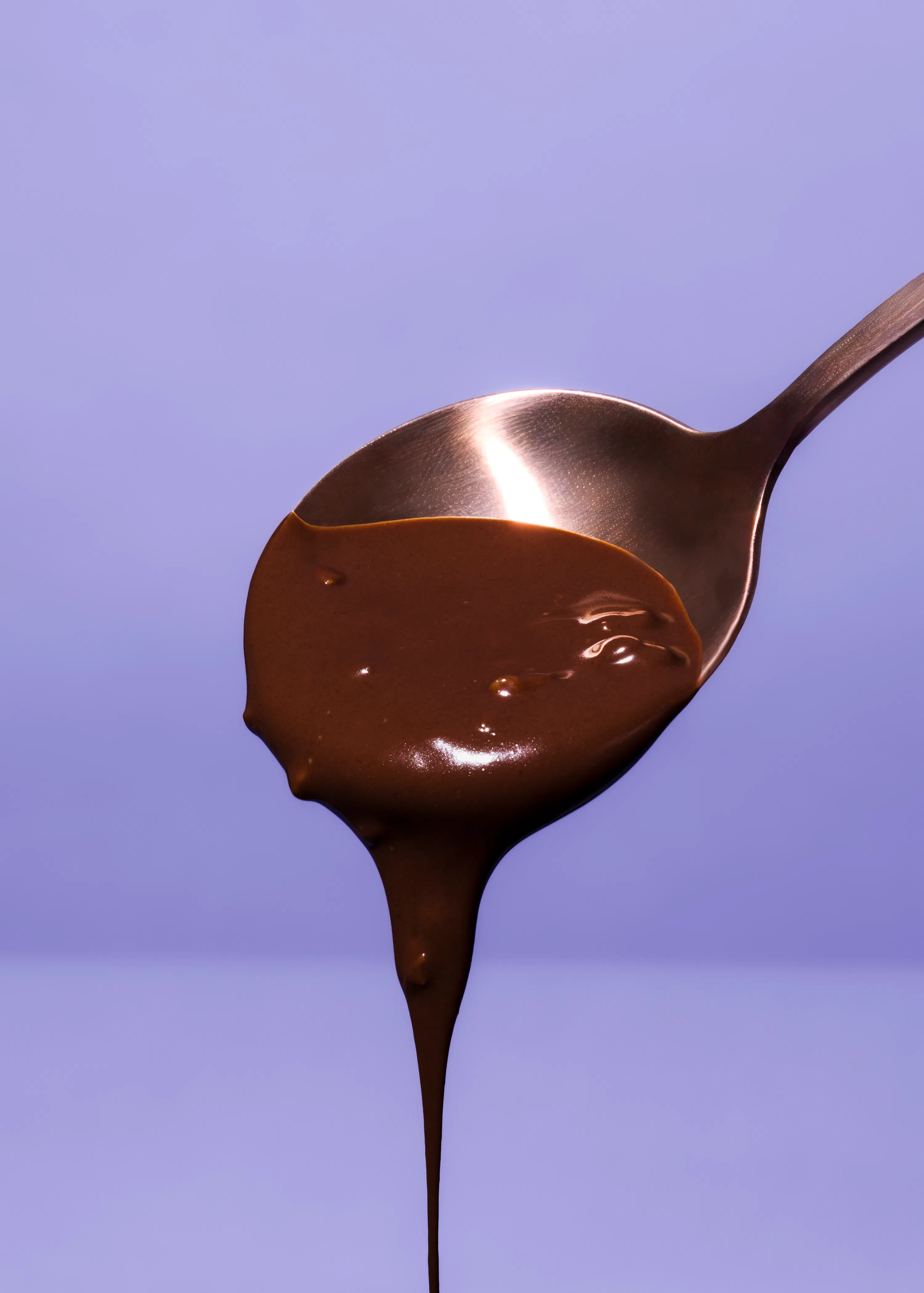 ein löffel mit geschmolzener schokolade, die vor einem hellvioletten hintergrund heruntertropft.