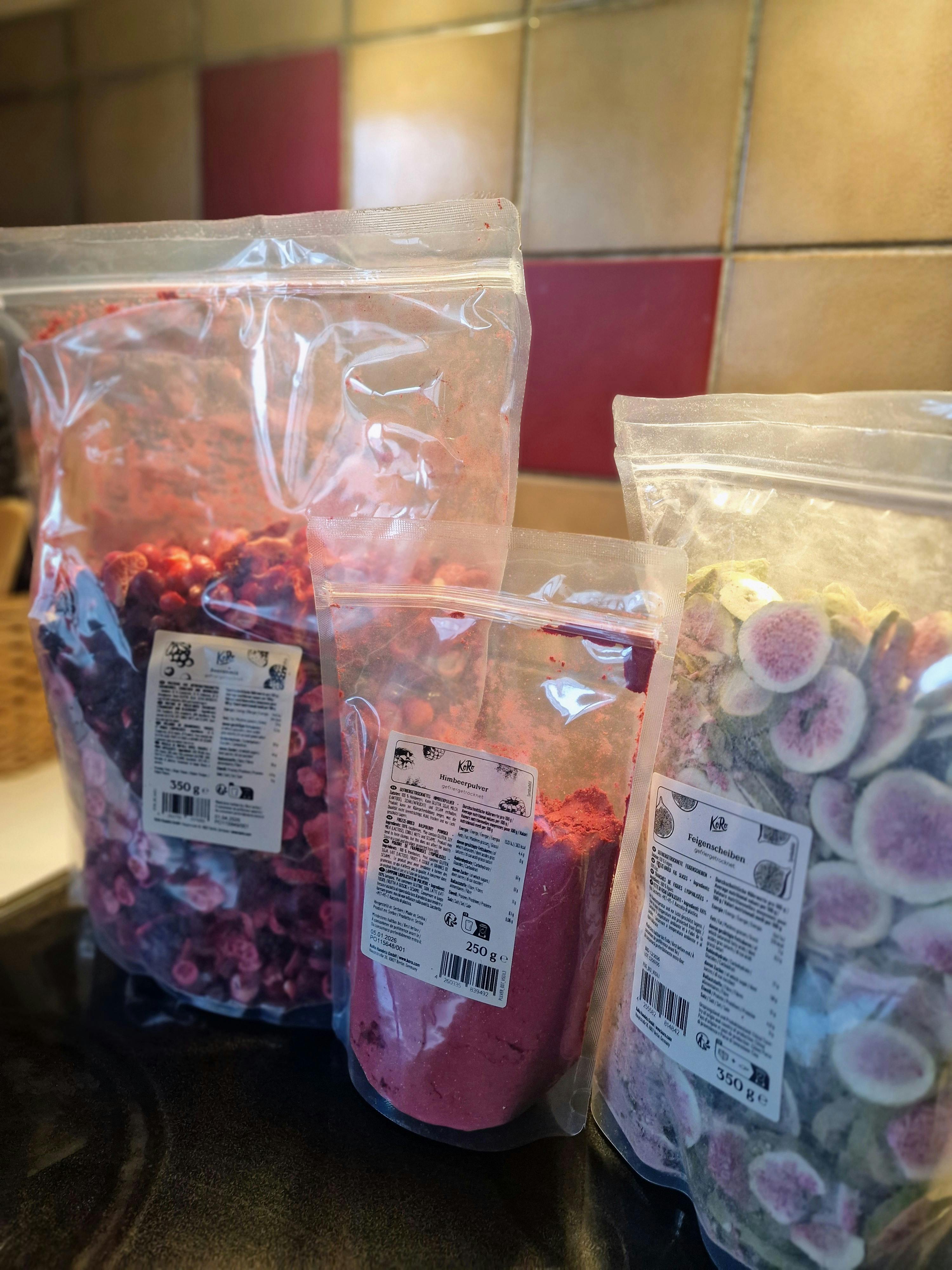 drei transparente beutel mit getrockneten früchten und himbeerpulver stehen auf einer theke vor einer gefliesten wand.