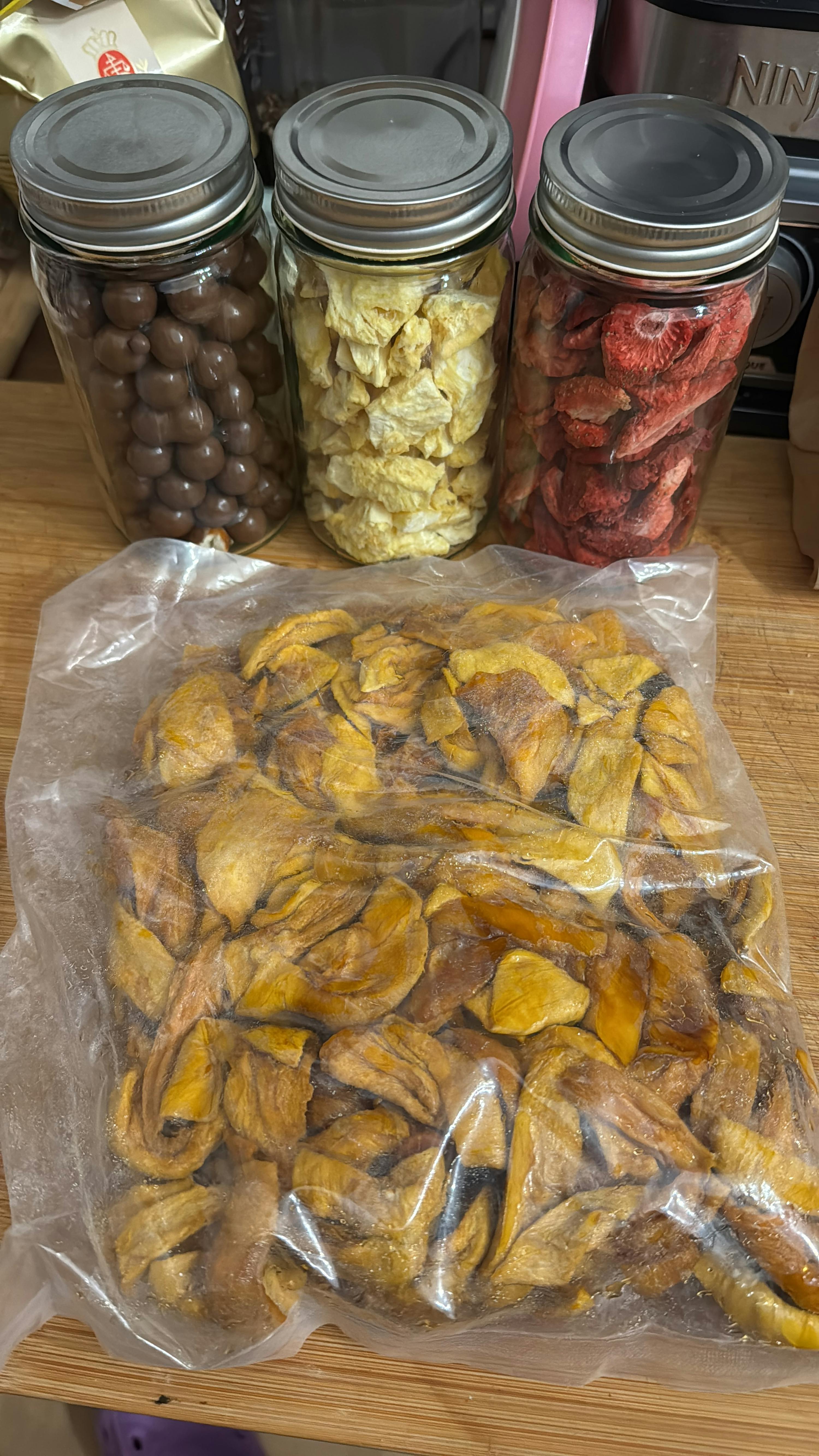 drei gläser mit gefriergetrockneten erdbeeren, ananas und schokoladenkugeln stehen neben einem beutel mit getrockneten mangoscheiben auf einem holzbrett.