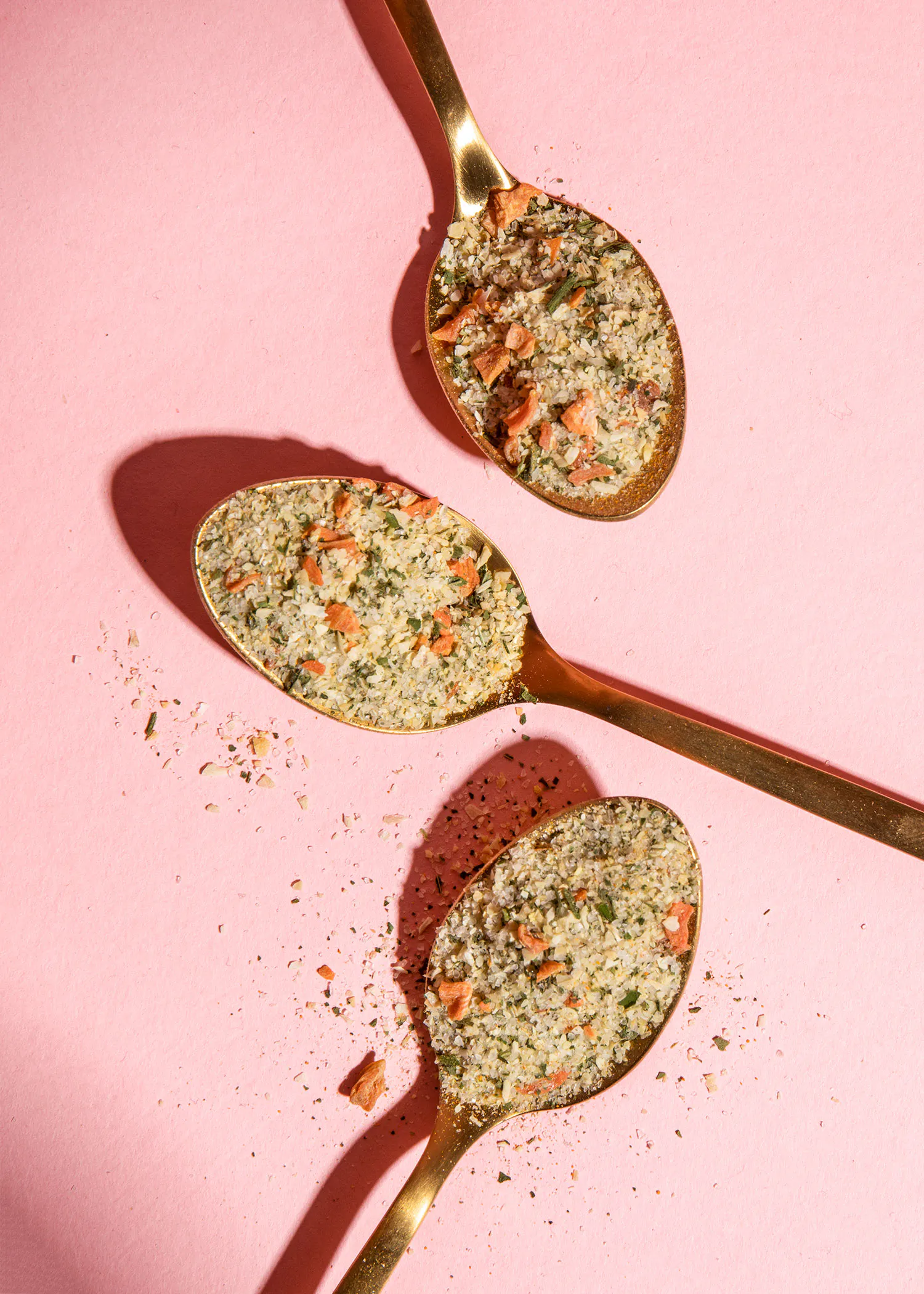 drei goldene Löffel mit Gemüse und Gewürzen auf einem rosa Hintergrund